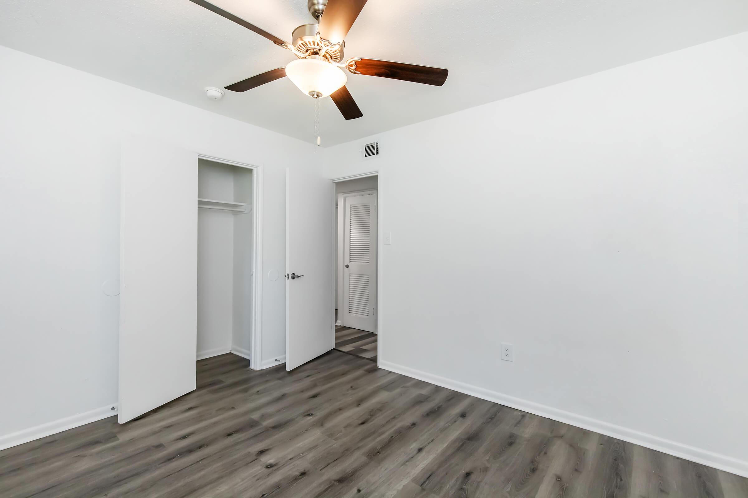 A well-lit room featuring a ceiling fan, light-colored walls, and wood-style flooring. Two white doors are visible: one leading to a closet and another to a hallway. The overall ambiance is clean and modern, suitable for a bedroom or living space.