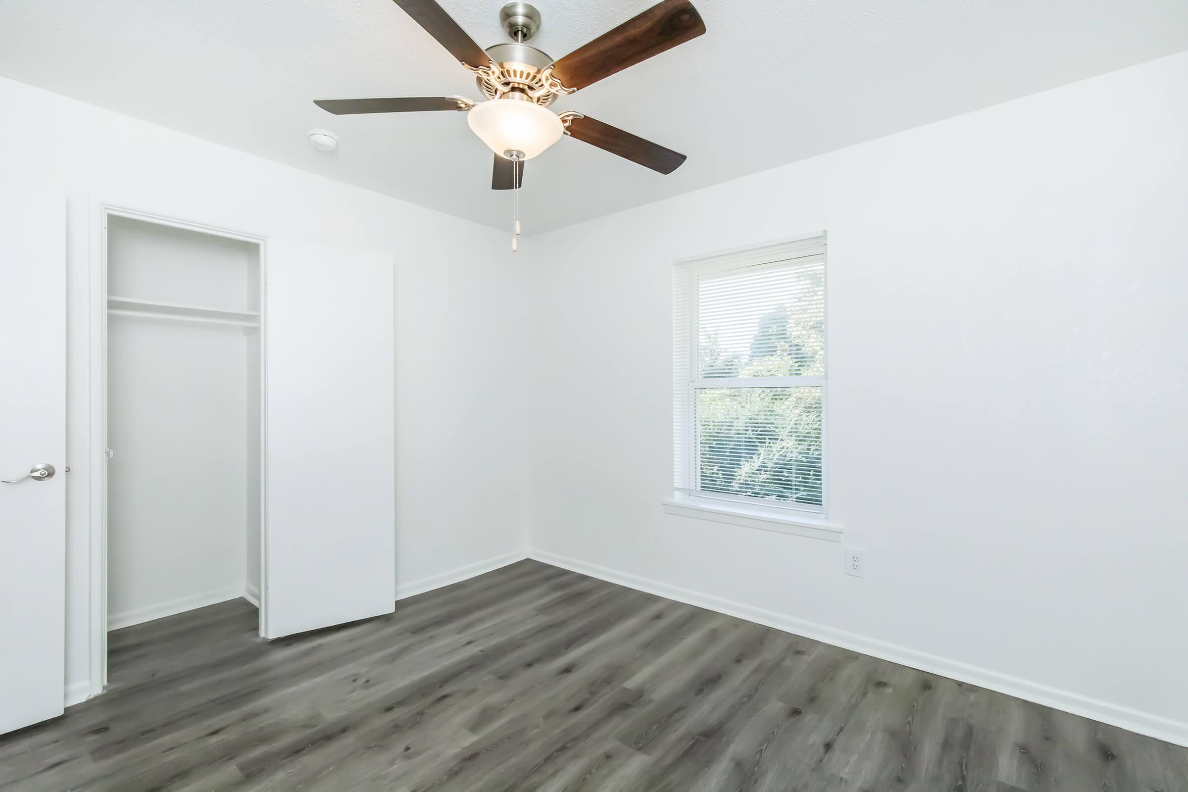A clean, empty room featuring a ceiling fan and a window allowing natural light. The walls are painted white, and there is a closet with double doors on one side. The flooring is a light wood laminate, creating a bright and airy atmosphere.