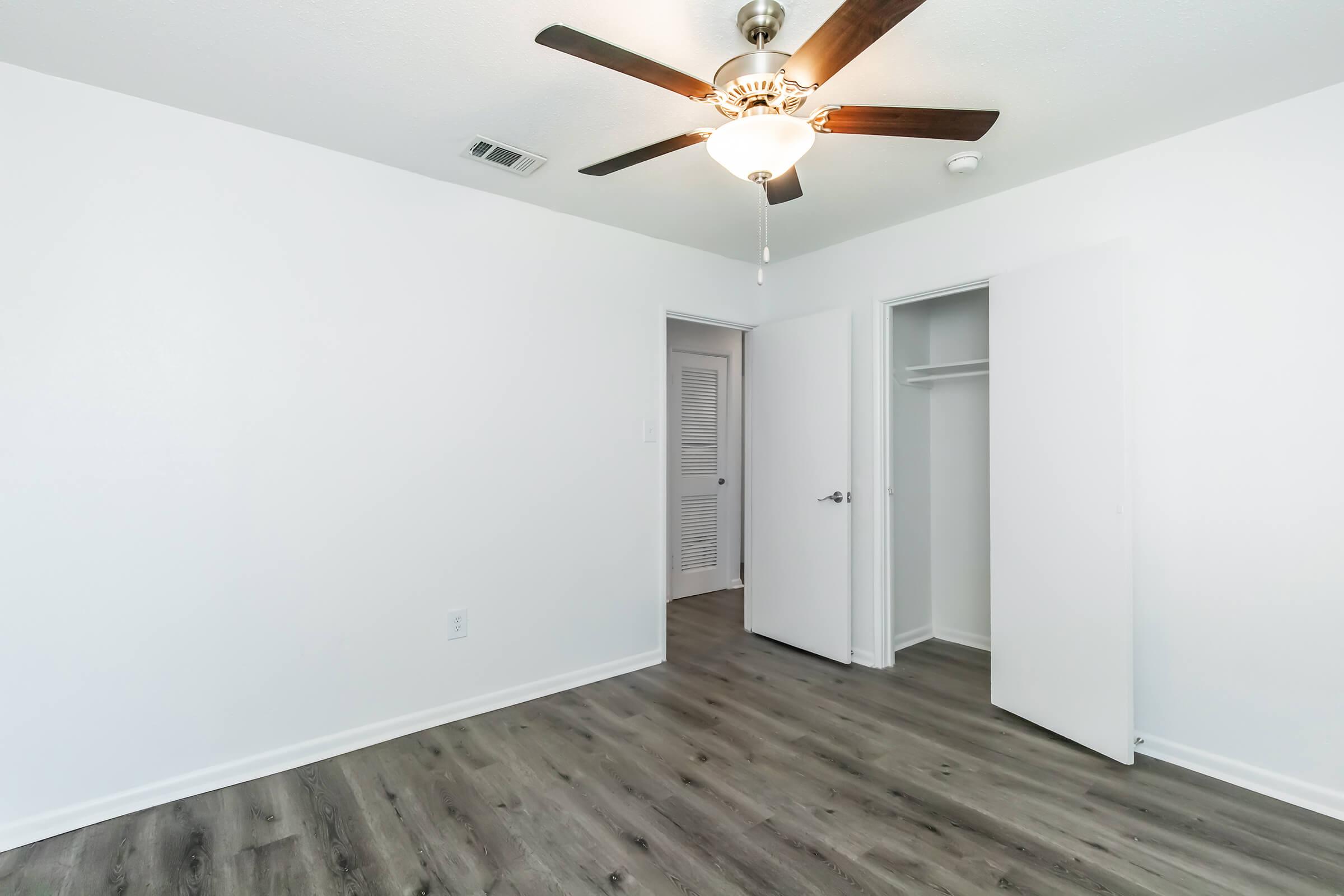 A bright, empty bedroom featuring a ceiling fan, light fixture, and white walls. There are two doors leading to a closet and another room. The floor is wood-like laminate, giving the space a modern feel. The room is uncluttered, ideal for personalization.