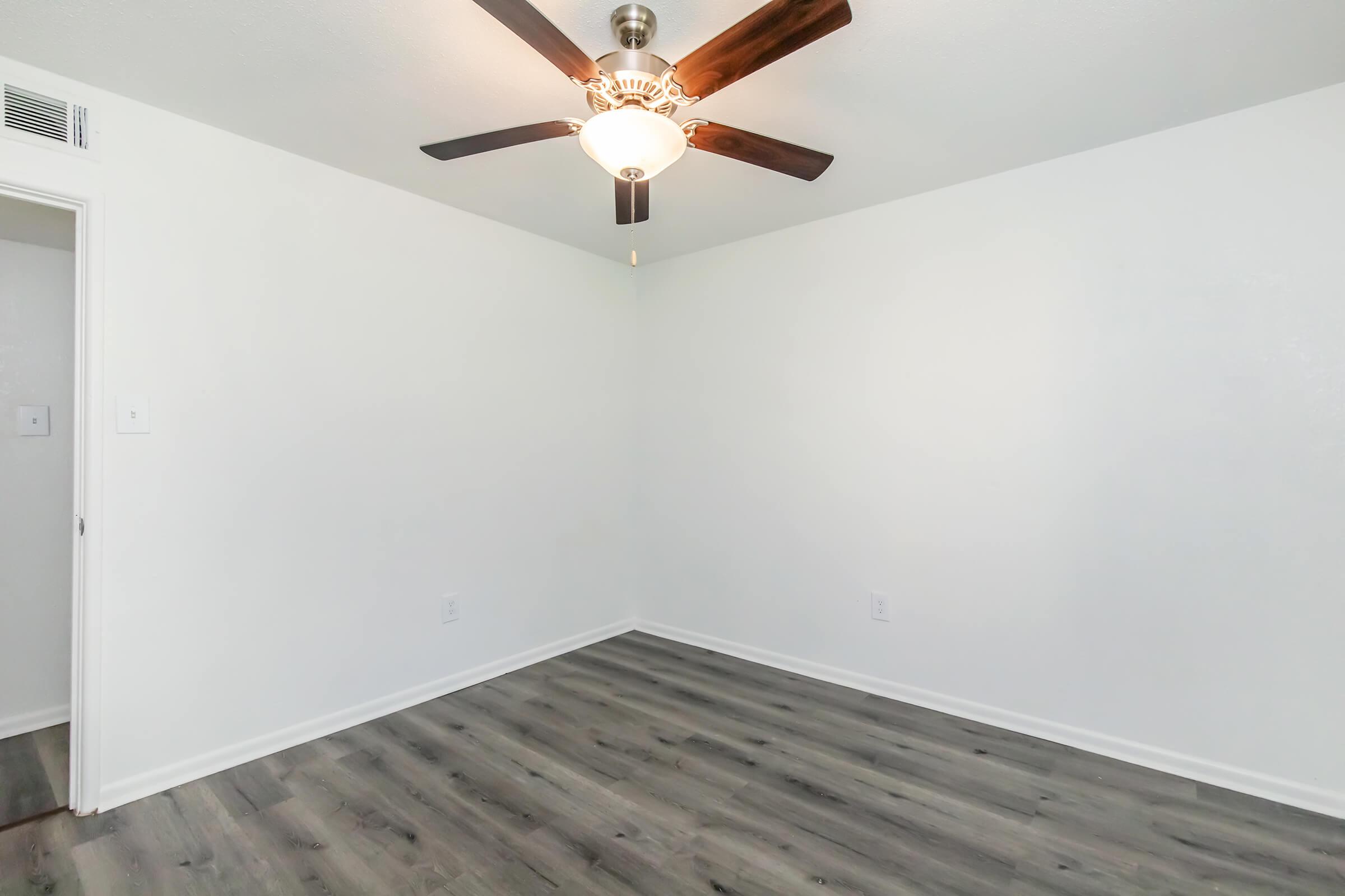 A clean, empty room with light gray walls and dark wood flooring. A ceiling fan with wooden blades and a light fixture is centered on the ceiling. There is a doorway leading to another area, and the room is well-lit with natural light.