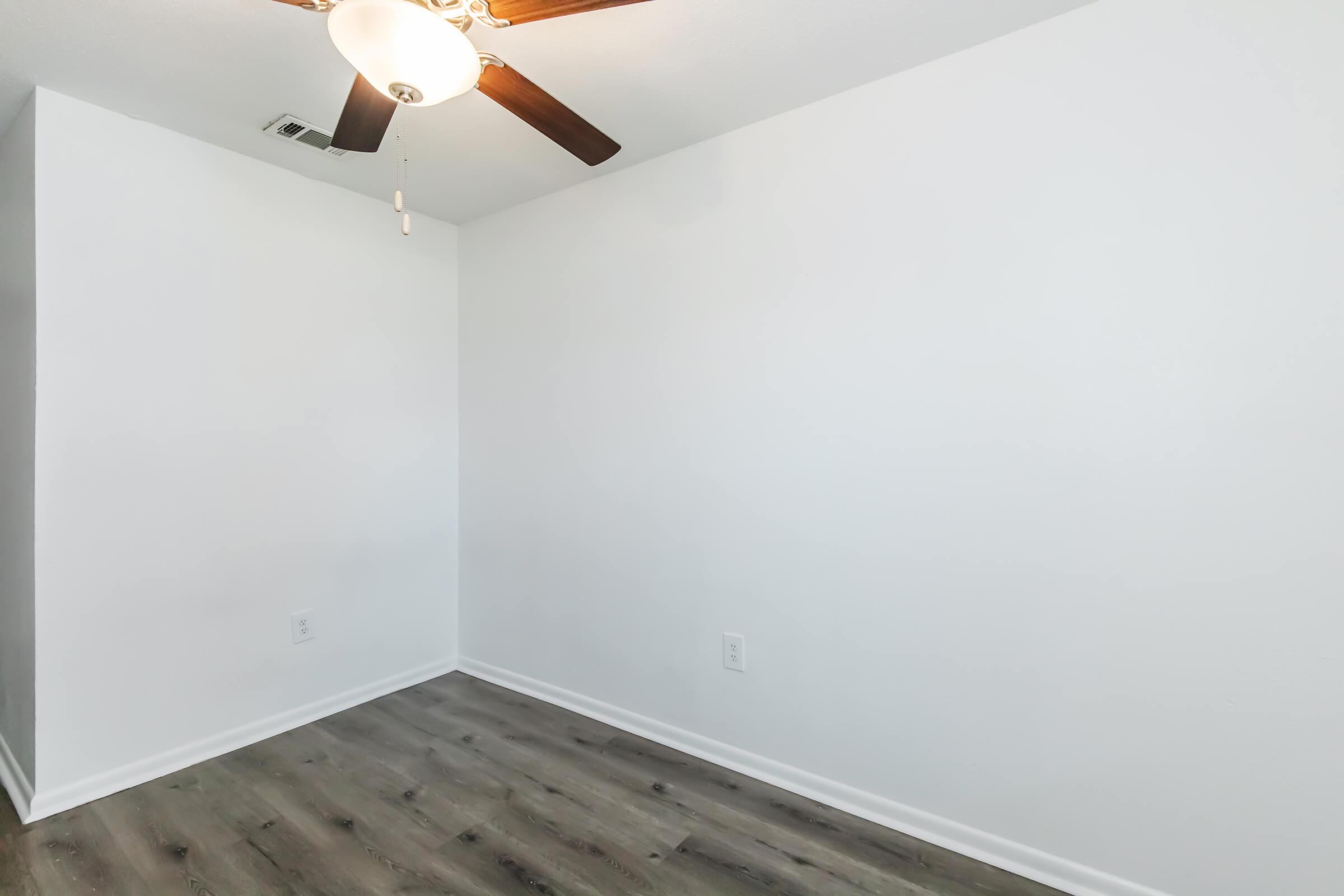 A bright, empty room with white walls and a ceiling fan. The flooring is a light wood laminate, and there are no furniture or decorations present, creating a clean and spacious atmosphere.
