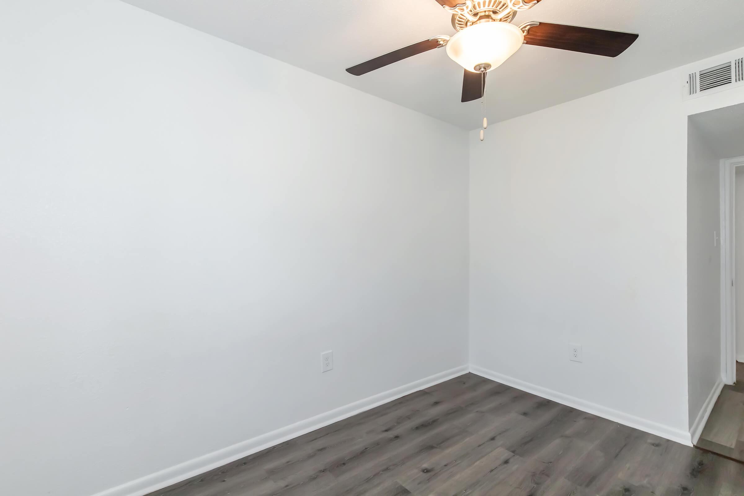 A bright, empty room with white walls and a ceiling fan. The floor is covered in a sleek, dark laminate. A doorway is visible on the right side, leading to another space, but the room is otherwise unfurnished and spacious, giving it a clean and airy feel.