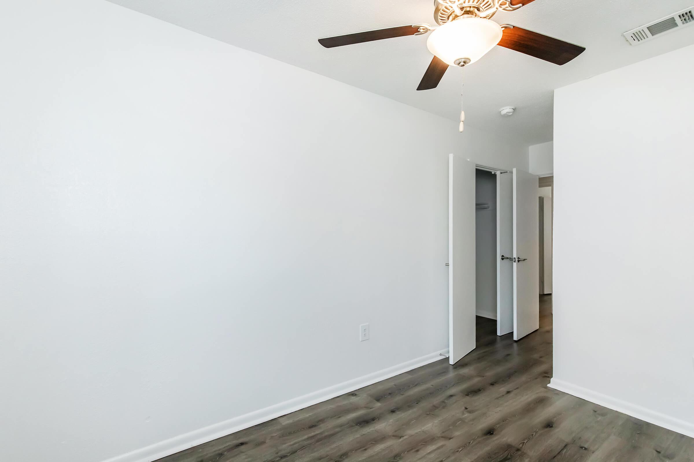 A bright, empty room with white walls and a ceiling fan. The floor features dark wood-like laminate, and there are two open doors leading to adjoining spaces. The overall atmosphere is clean and modern, suitable for various uses.