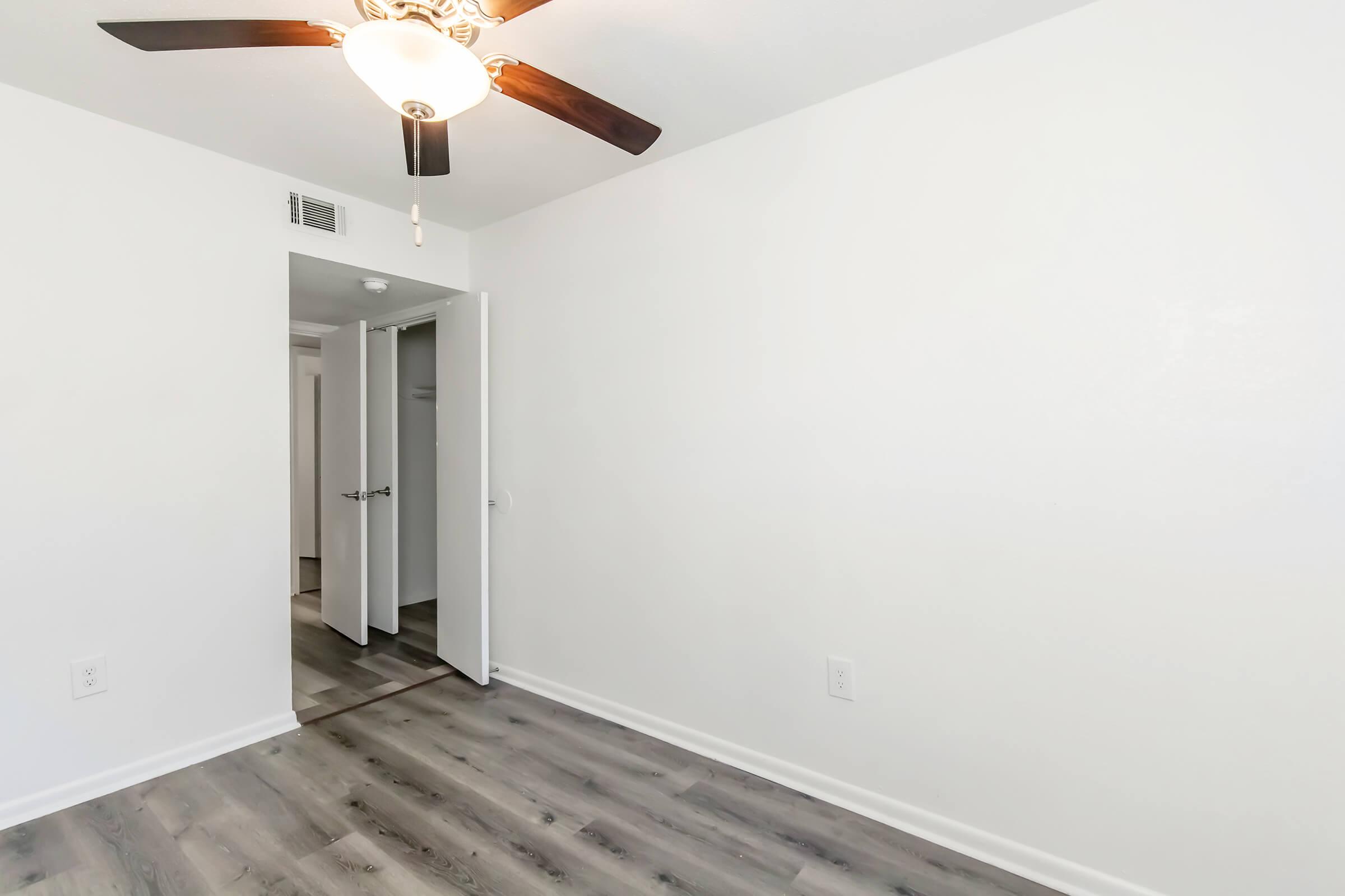 A well-lit room with light-colored walls and a ceiling fan. The floor has dark laminate wood. There is a partially open door leading to another room, and the space appears clean and uncluttered.