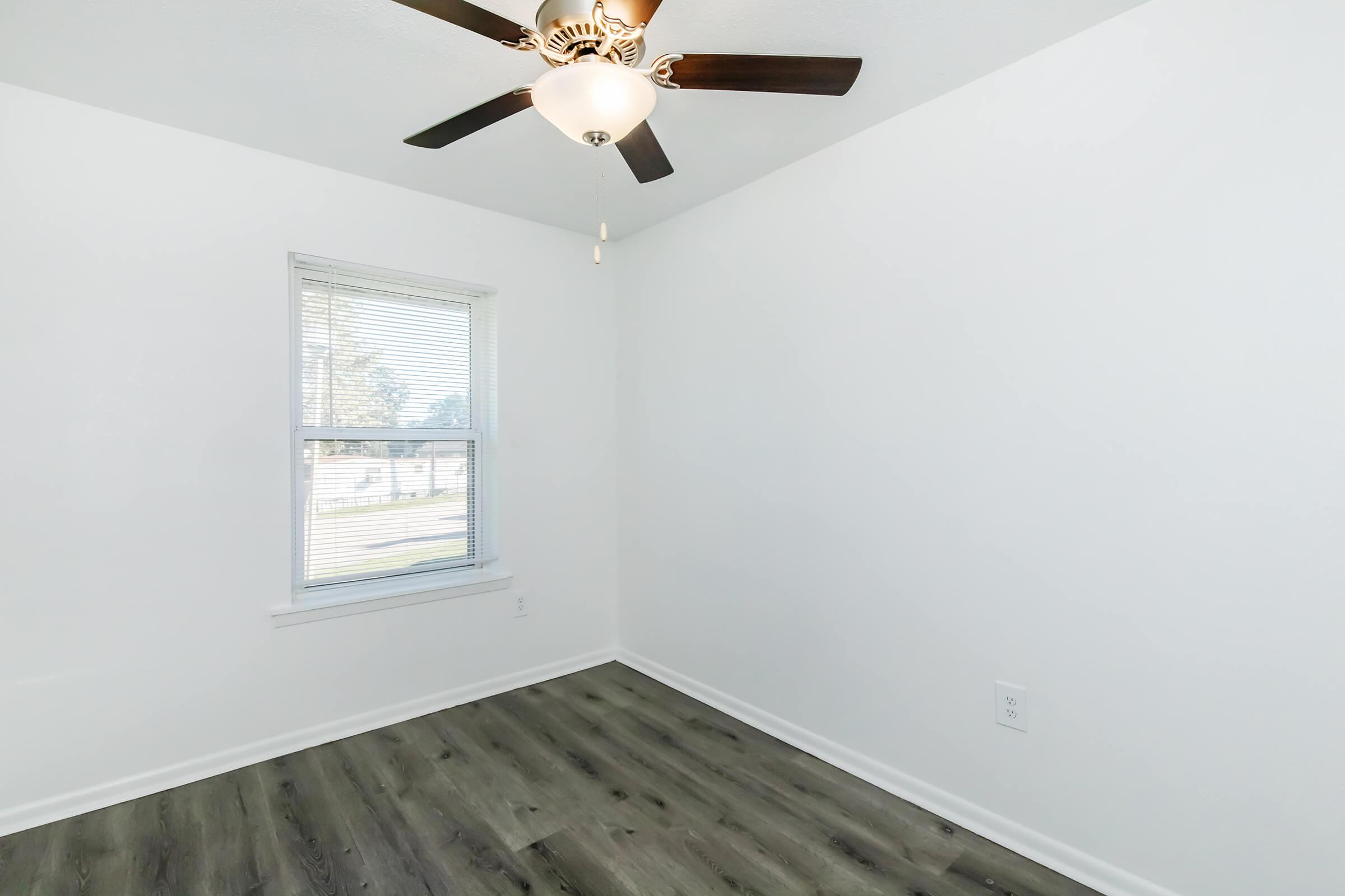 A well-lit room with a ceiling fan and a window, featuring light-colored walls and a wood-like floor. The space appears empty and clean, suitable for various uses such as a bedroom or office.