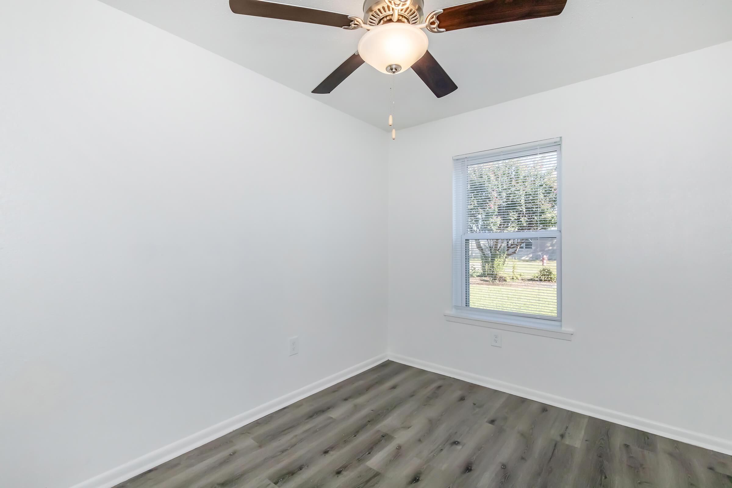 A well-lit, empty room with light-colored walls, a ceiling fan with a light fixture, and a window revealing greenery outside. The floor features light wood laminate flooring. This space appears to be ready for furniture or decoration.