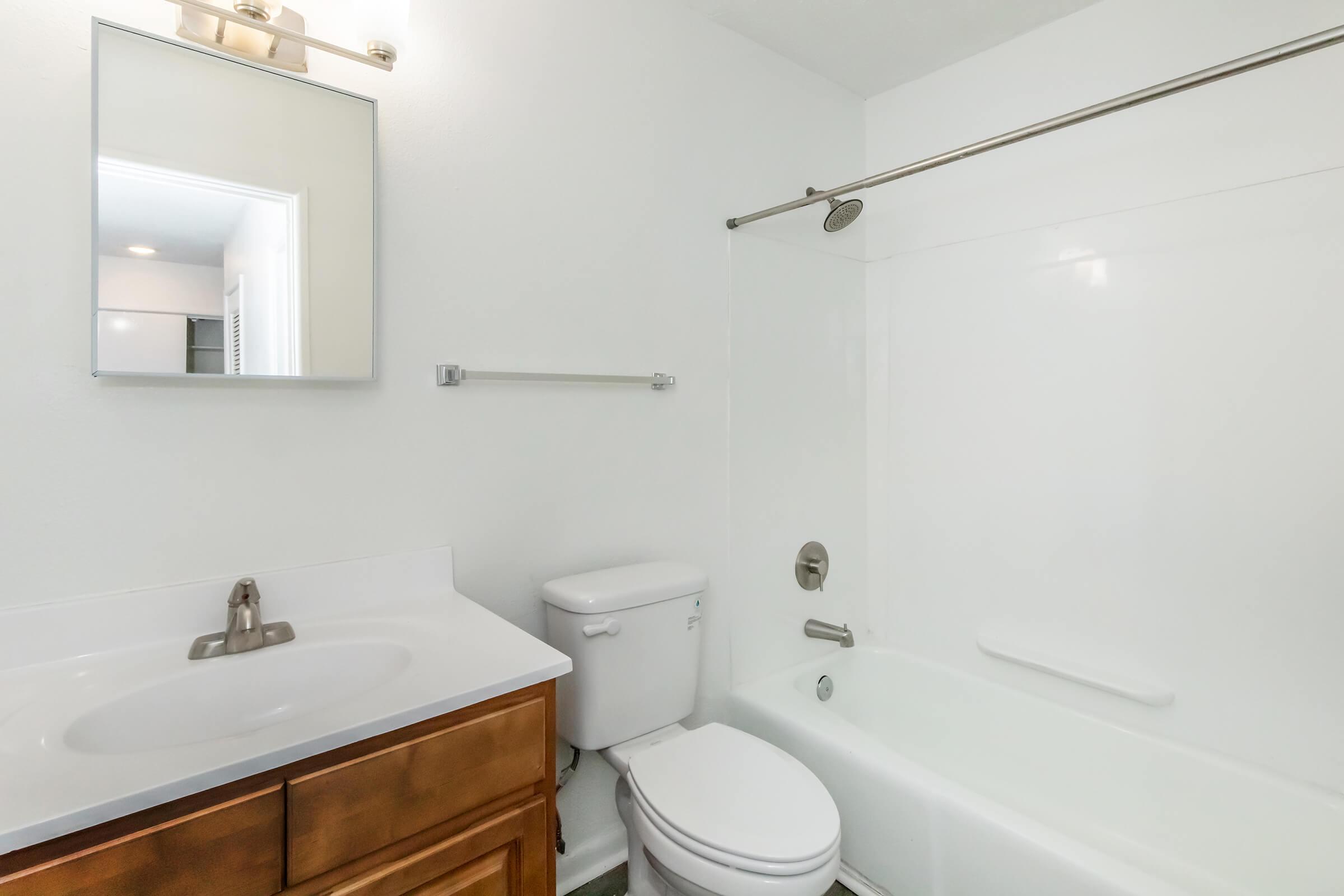 A clean, modern bathroom featuring a white bathtub with a shower, a vanity with a sink and wooden cabinetry, a toilet, a wall-mounted mirror, and a towel rack. The walls and fixtures are primarily white, creating a bright and spacious appearance.