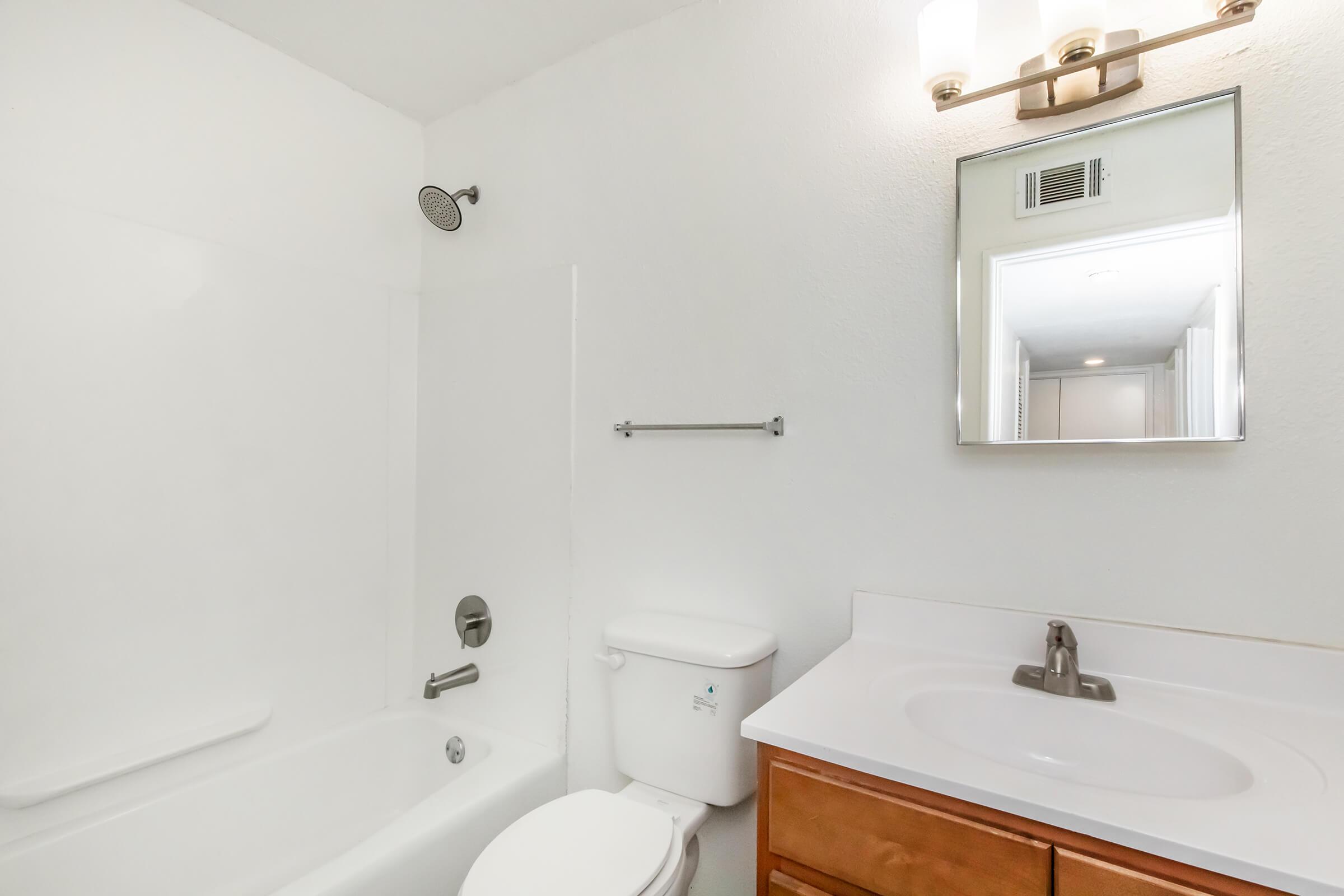 A clean, modern bathroom featuring a white bathtub with a shower head, a toilet, and a sink with a wooden cabinet. The walls are painted white, and there is a mirror above the sink, with a light fixture mounted above it. The space appears bright and well-maintained.