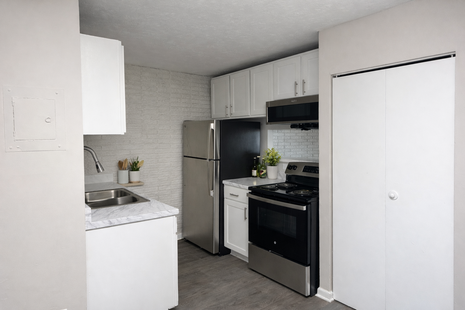 Modern kitchen featuring white cabinetry, a stainless steel refrigerator, and an electric stove. The countertop is a light marble finish, and there's a small sink. A white brick wall adds texture, while a sliding door offers access to another room or storage. Fresh herbs in pots add a touch of greenery.