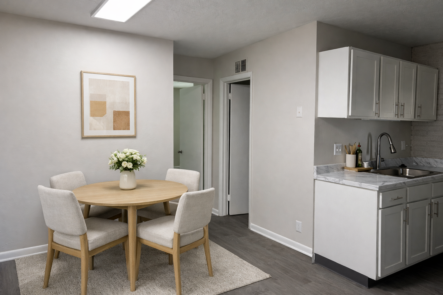 A modern kitchen and dining area featuring a round wooden table with four upholstered chairs. A vase of white flowers sits on the table. The kitchen has light-colored cabinets and a marble countertop, with a doorway leading to another room. The walls are painted in neutral tones, creating a bright and inviting atmosphere.