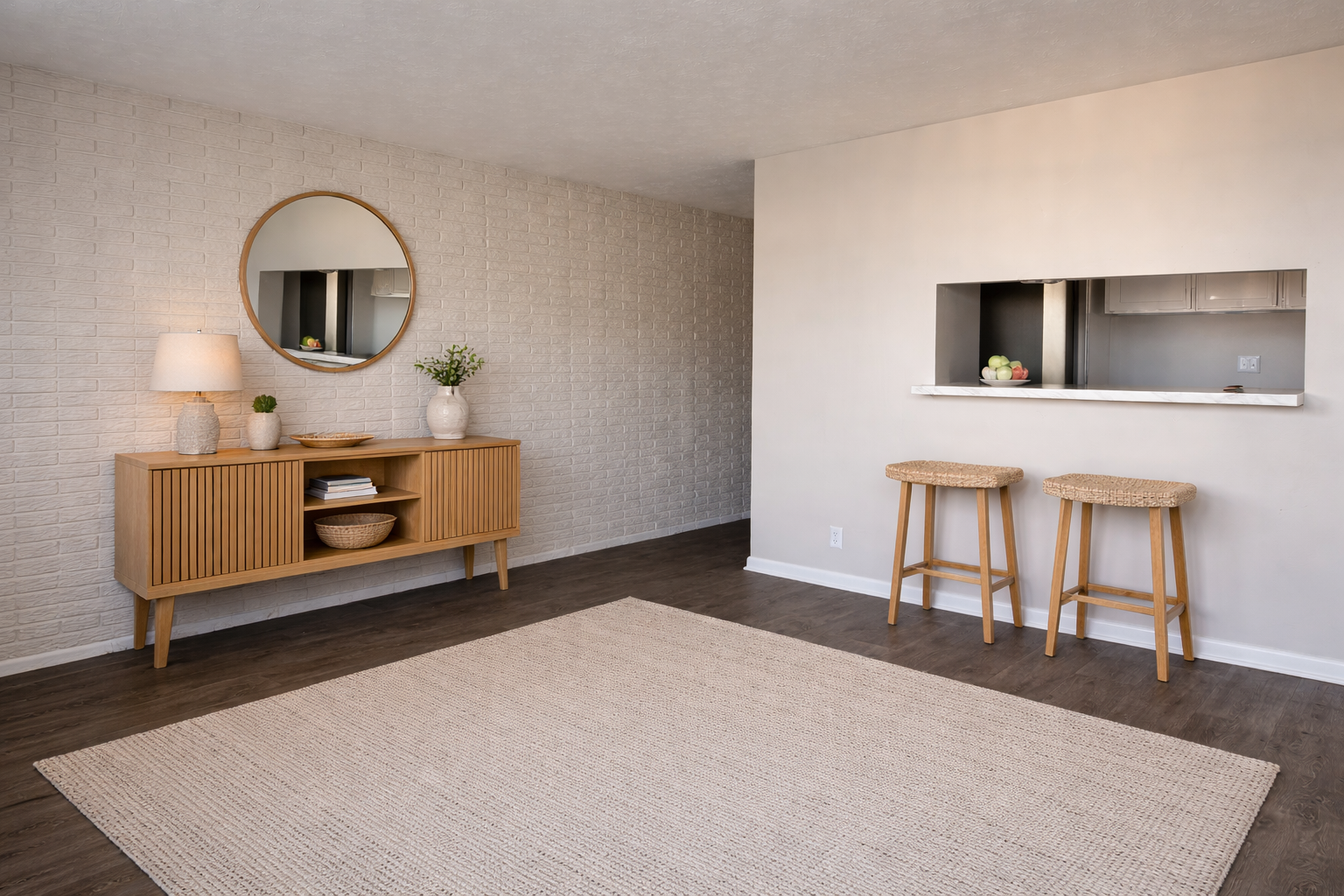 A modern, bright living space featuring a light-colored wall with a round mirror, a wooden console table with a lamp and decorative plant, and a woven rug. Adjacent to the seating area are two simple bar stools at a white countertop with a view of an open kitchen in the background.