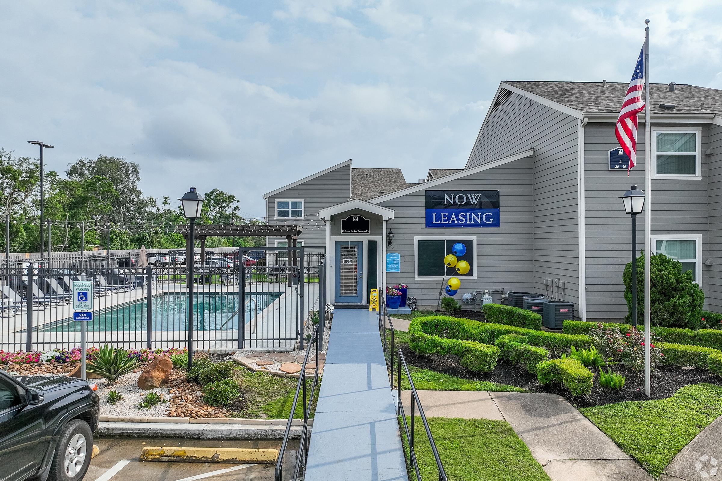 A leasing office with a blue ramp leading to the entrance. The building features a large "NOW LEASING" sign, surrounded by well-maintained landscaping including shrubs and flowers. In the background, there's a swimming pool area enclosed by a fence, and an American flag is visible.