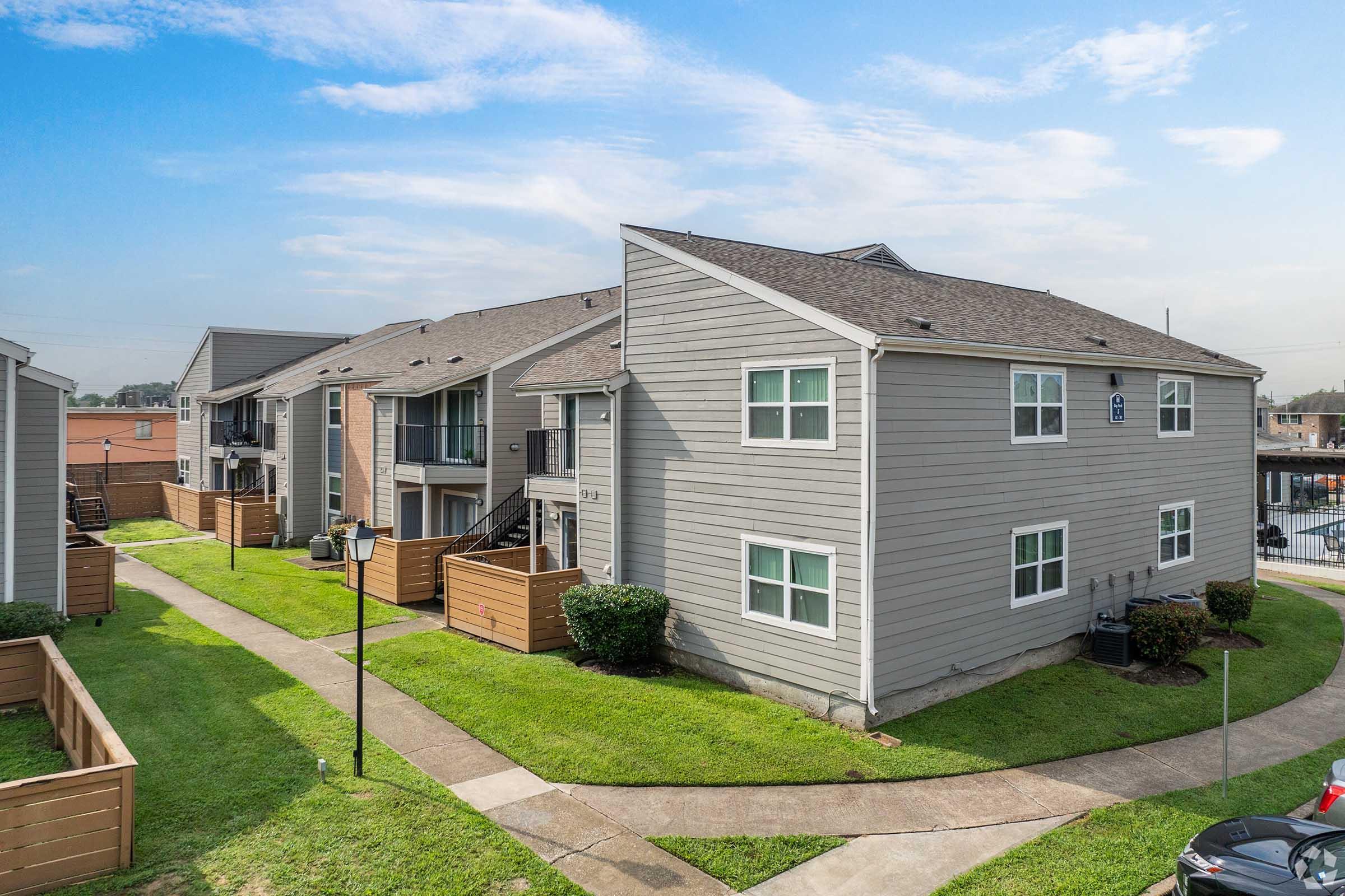 A residential complex featuring two-story buildings with gray siding. The area includes well-maintained lawns, pathways, and decorative lighting. Several units have balconies, and the layout is designed for easy access. The sky is clear with a few scattered clouds.