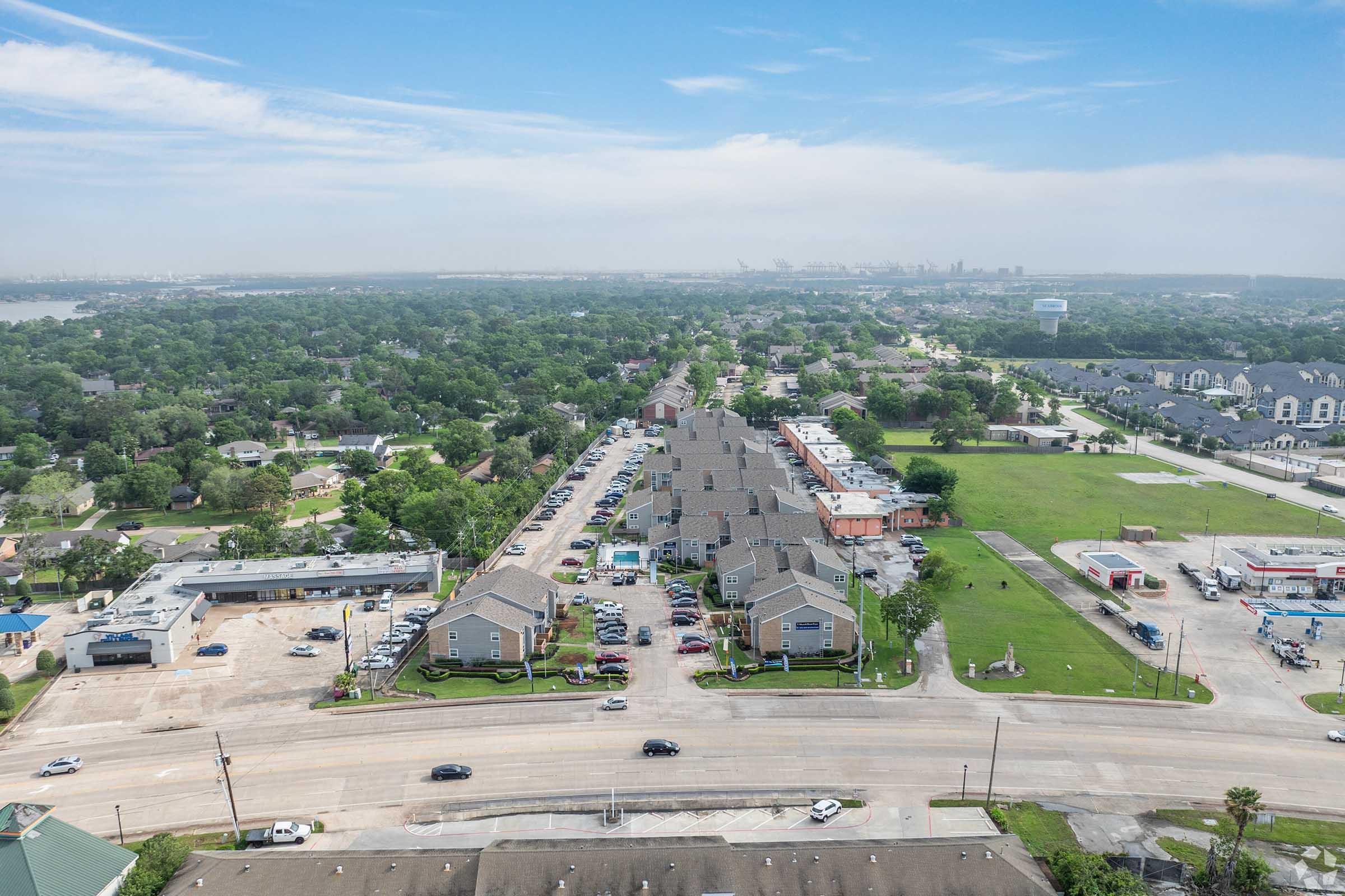 Aerial view of a suburban area featuring multiple residential buildings along a road, with parking lots and green spaces. In the background, the skyline of a city can be seen, along with open fields and nearby commercial establishments. The scene shows a blend of urban and natural landscapes under a clear blue sky.