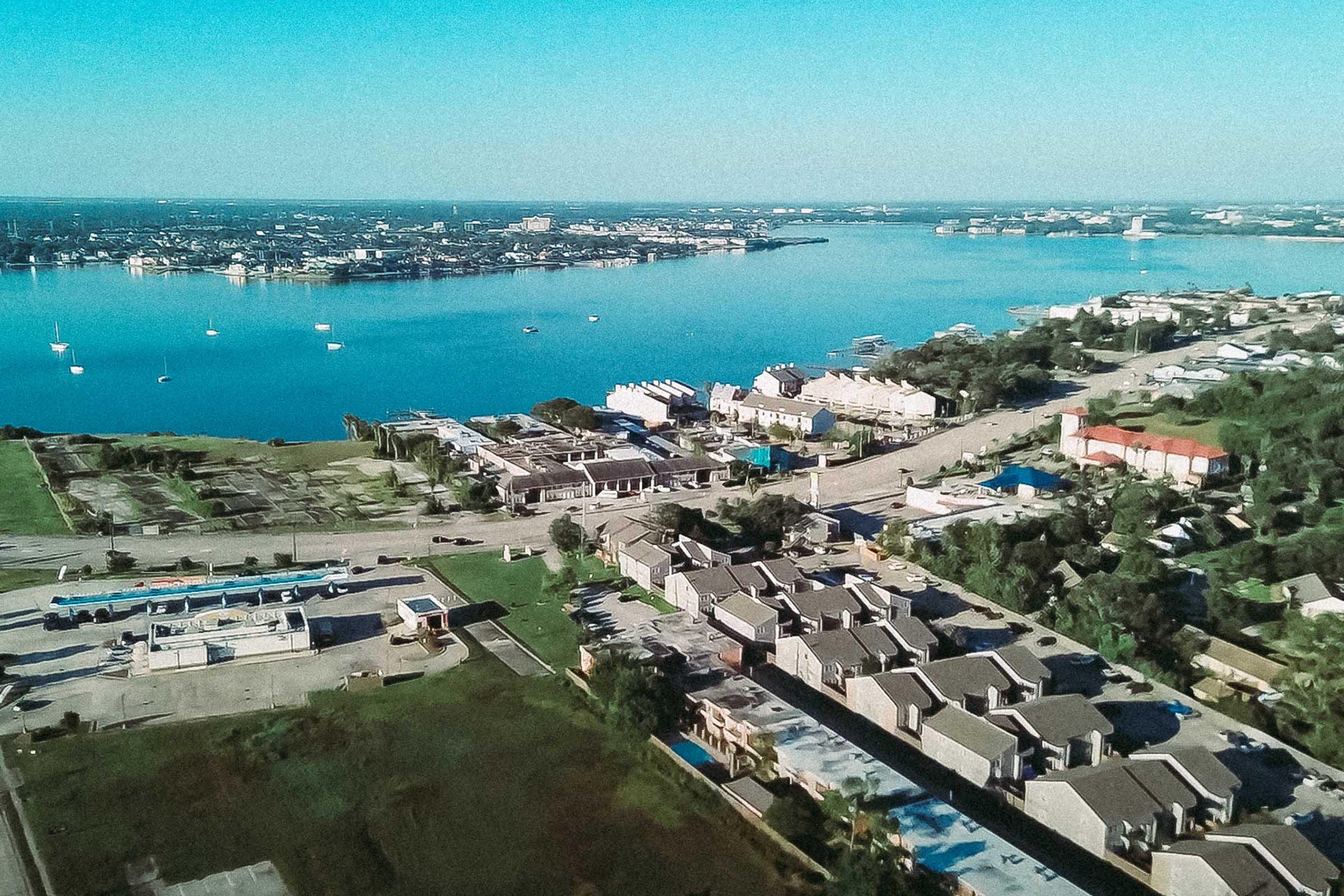 Aerial view of a coastal area featuring residential buildings along the shore, a marina with boats, and calm blue waters. The landscape includes green spaces and a road, under clear blue skies with distant land visible on the horizon.