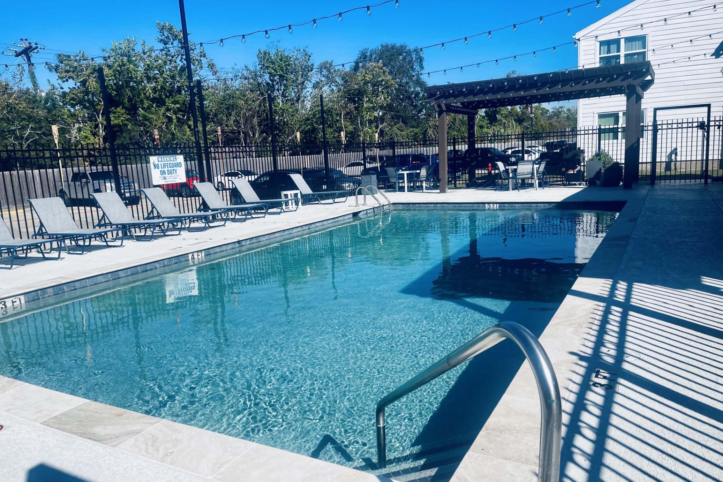 A clean swimming pool with crystal-clear water, surrounded by lounge chairs. The pool area features a shaded pergola and is enclosed by a black fence. Soft lighting strings are hung above, and lush green trees are visible in the background. The scene is bright and inviting, ideal for relaxation.