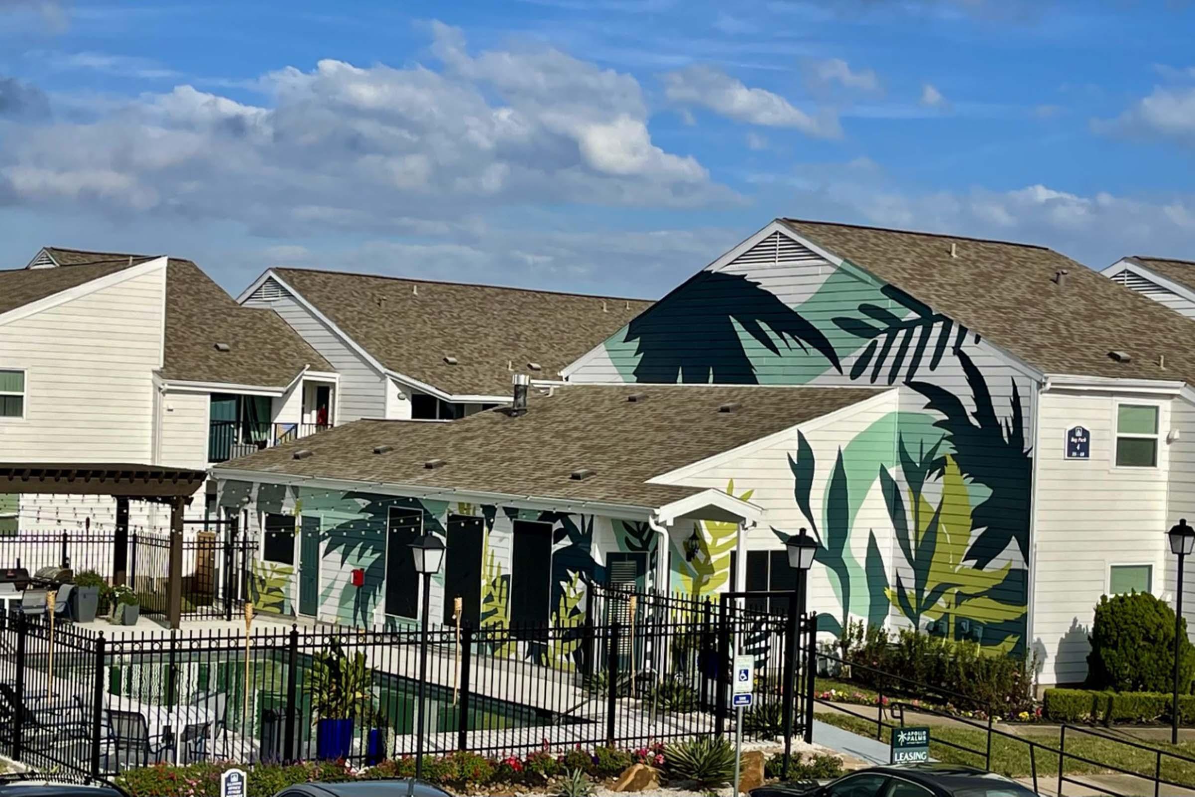 A view of a residential building featuring a large, tropical-themed mural on one side. The building is surrounded by a black fence, with a pool area visible in the foreground. The sky is partly cloudy, adding a bright atmosphere to the scene. Lush greenery and flower beds enhance the inviting appearance of the property.