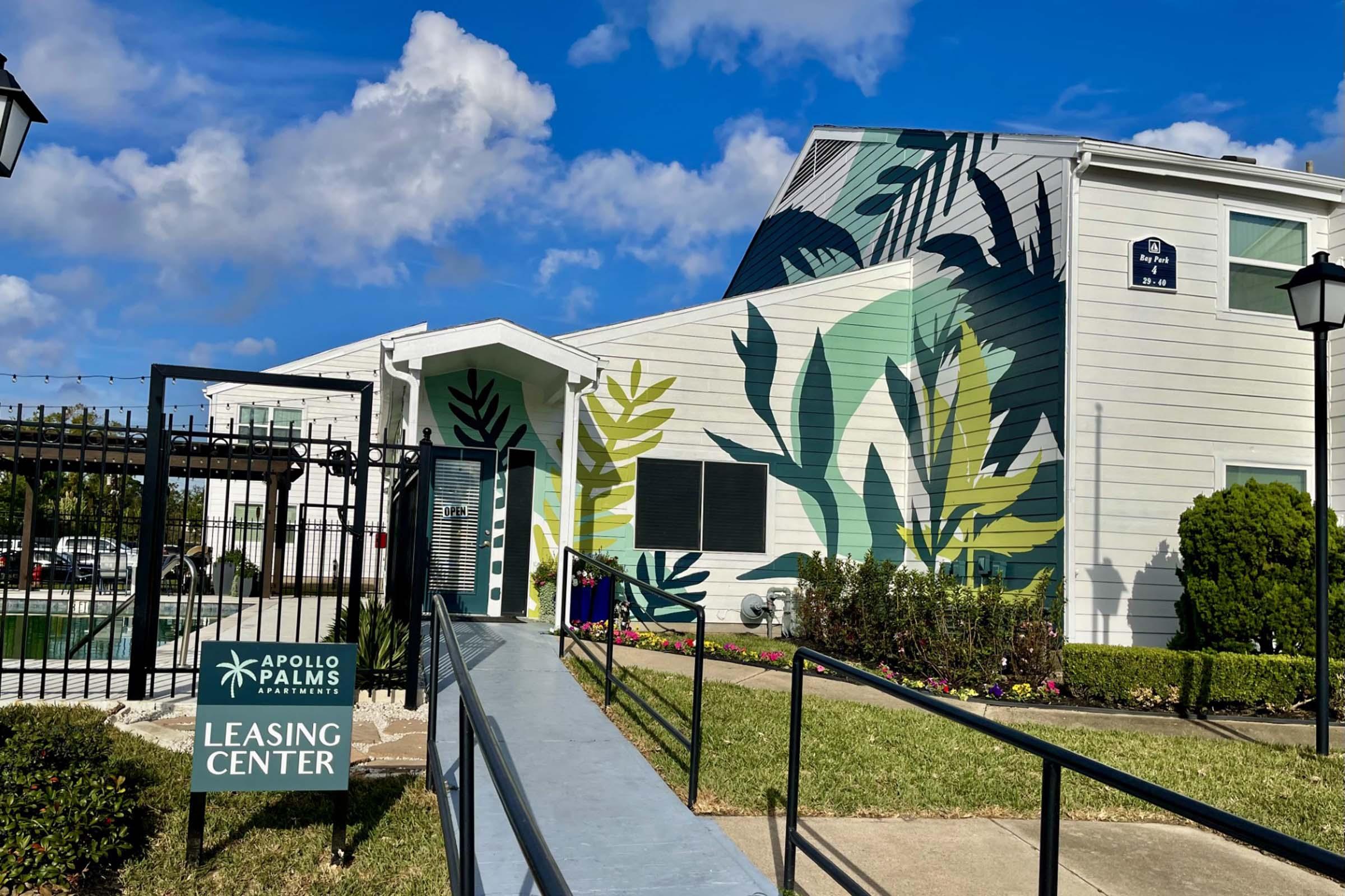 Leasing center building for Apollo Palms Apartments, featuring a colorful mural of tropical plants on one side. A walkway leads to the entrance, with green grass and flowers surrounding the area. The sky is partly cloudy, adding to the pleasant atmosphere.