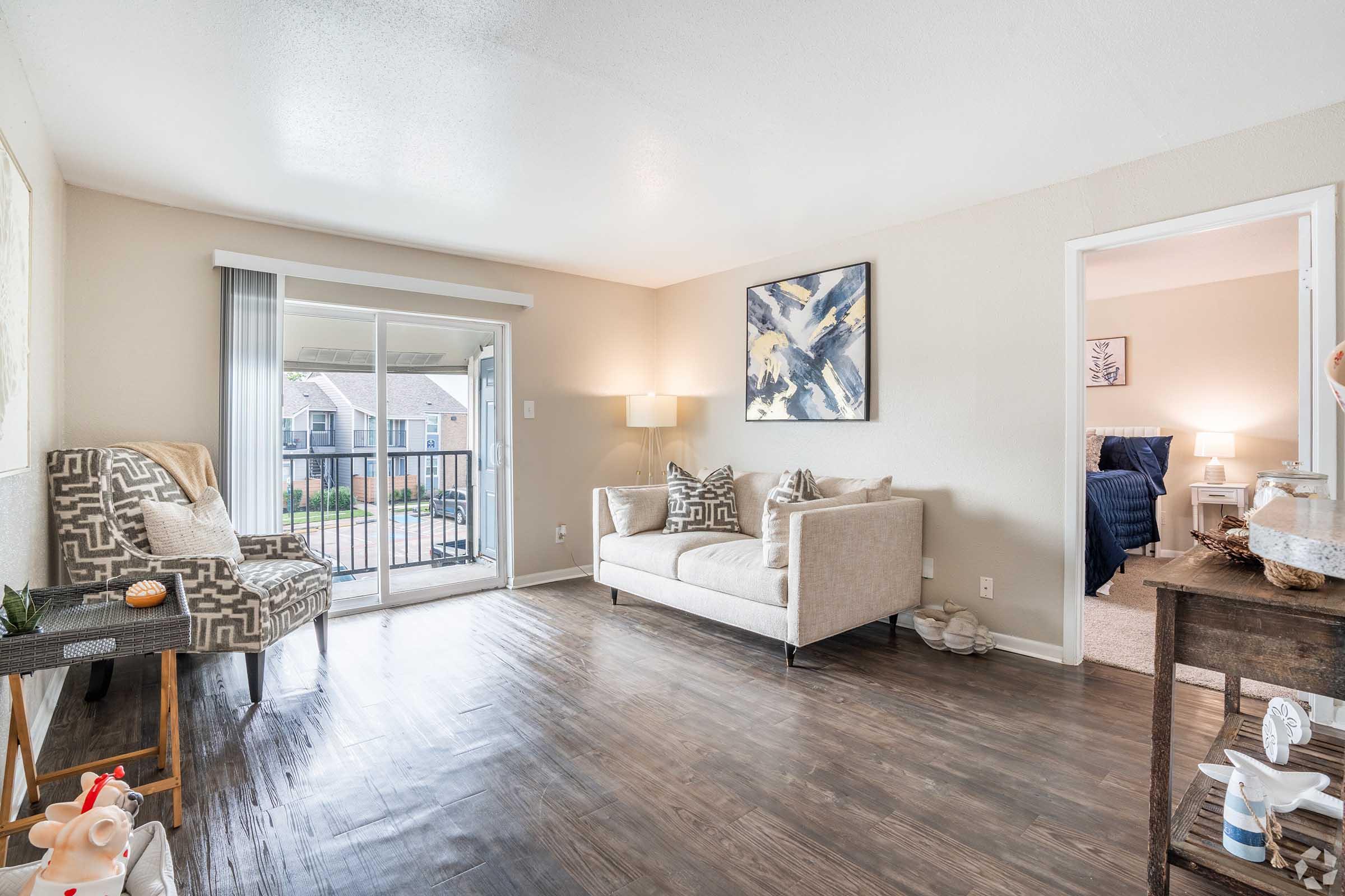 A bright, modern living room with hardwood flooring, featuring a beige sofa, patterned armchair, and decorative pillows. There is a sliding glass door leading to a balcony, and a small side table with decorative items. A painting hangs on the wall, adding a pop of color to the neutral decor.