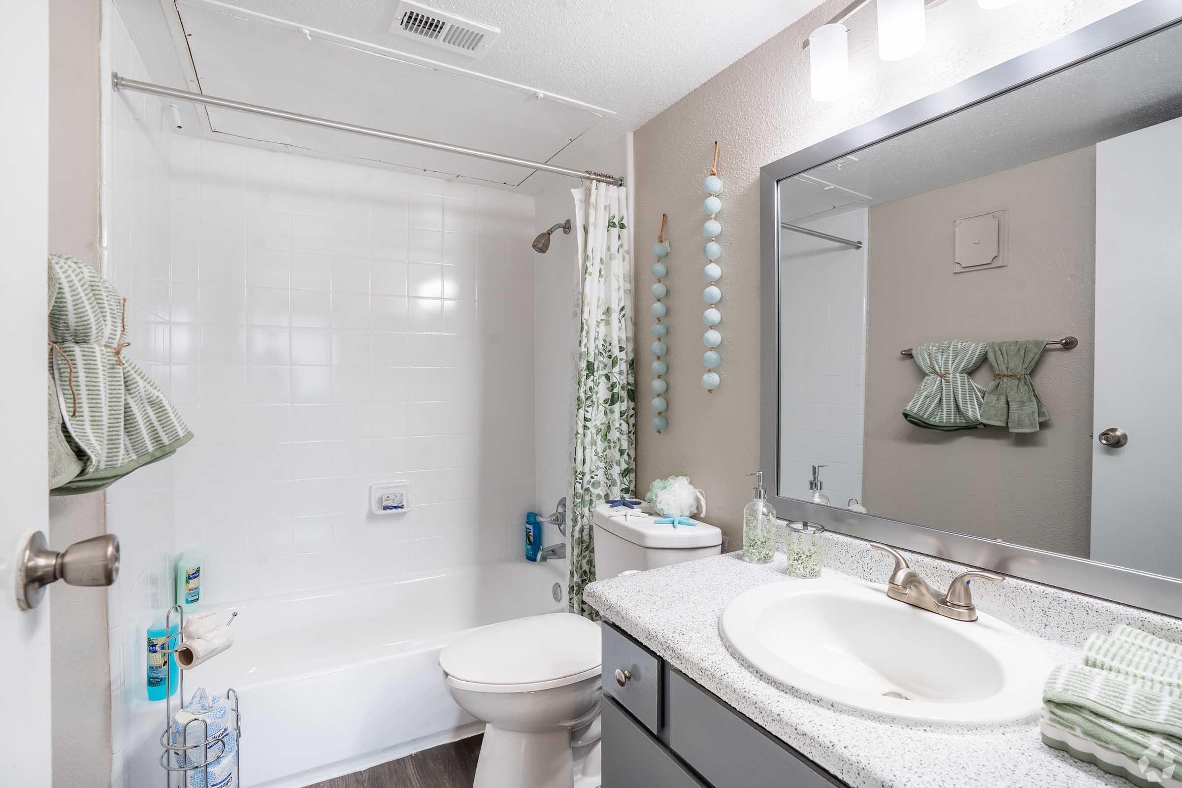 A clean and well-lit bathroom featuring a bathtub with a white shower curtain, a toilet, and a sink with a countertop. Decorative items include a green-themed towel set and hanging ornaments. The walls are painted in a neutral tone, enhancing the serene atmosphere of the space.