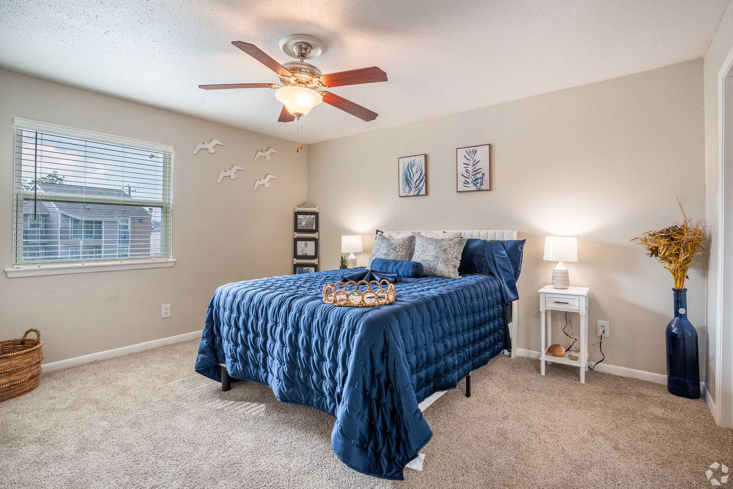 A cozy bedroom featuring a navy blue quilt on a bed with decorative pillows, a ceiling fan, and two lamps on nightstands. There’s a small table with a decorative tray and a woven basket. Wall art depicting nature themes and decorative accents add a personal touch. Natural light comes in through a window.