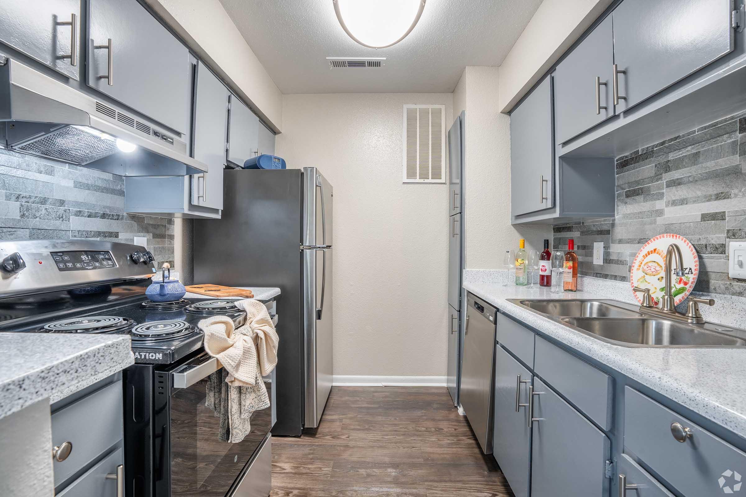 A modern kitchen featuring gray cabinetry, stainless steel appliances, and a silver refrigerator. The countertop is light-colored with a stovetop and oven. A small window allows natural light, and there are decorative items on the counter. The flooring is dark wood, creating a stylish contrast.