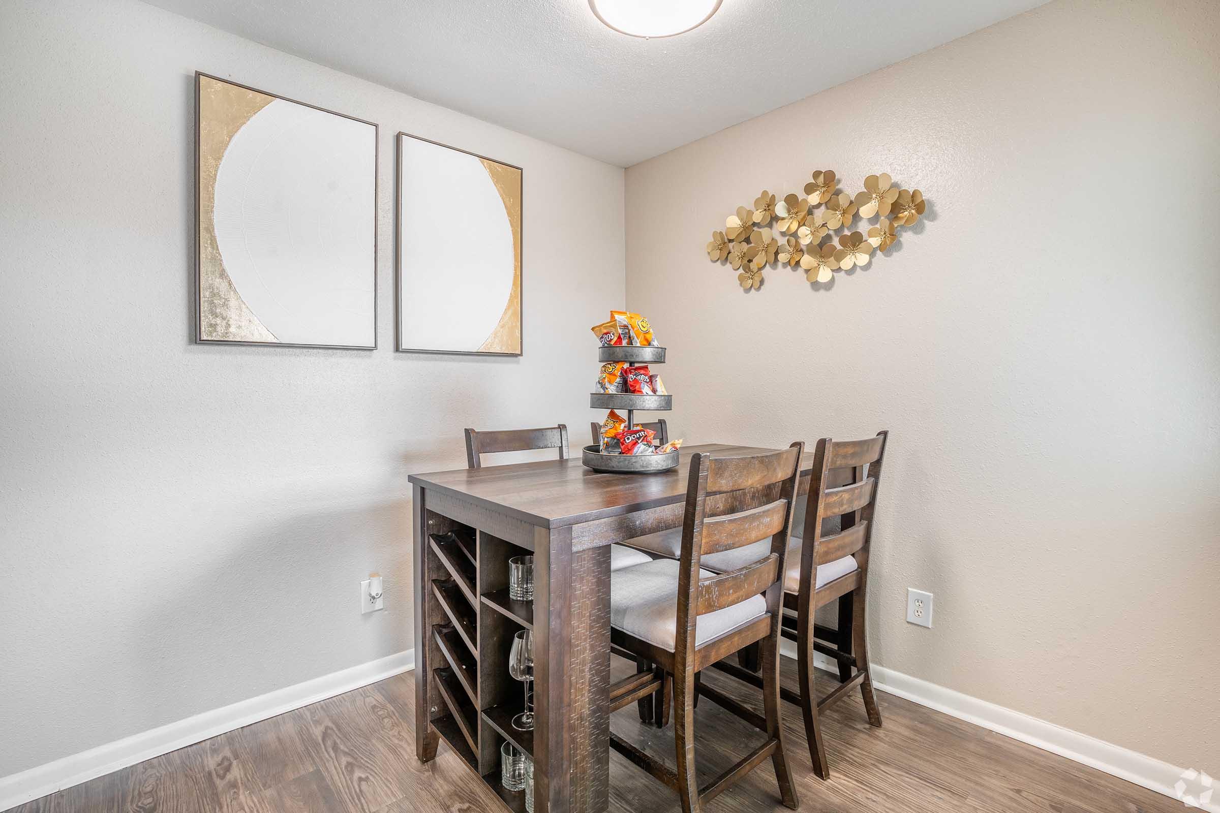 A modern dining area featuring a wooden table with four chairs, a tiered snack stand, and decorative wall art. The walls are painted in a neutral tone, and two circular framed artworks are displayed above the table. The space is well-lit and tidy, creating a welcoming atmosphere.