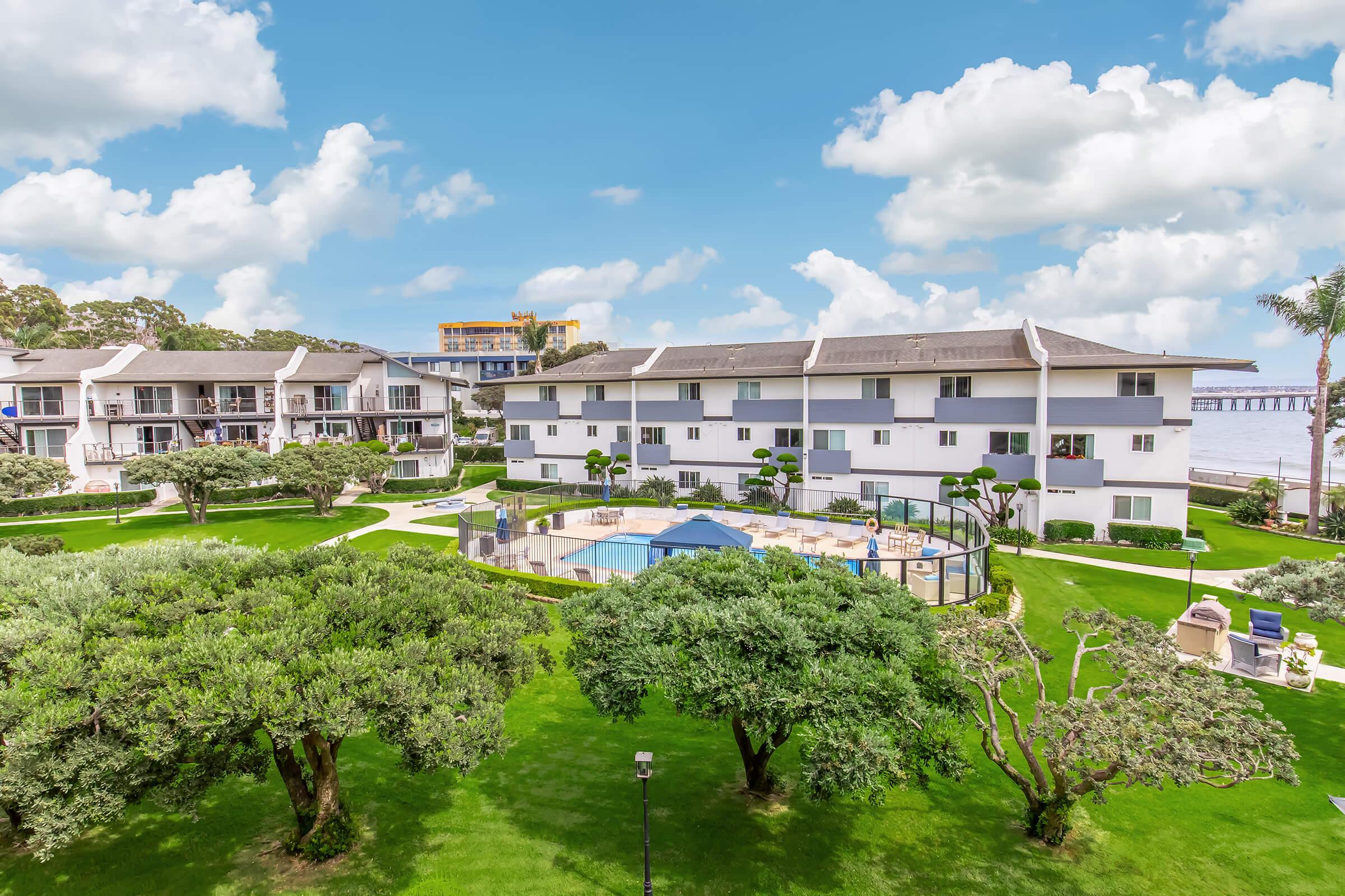A scenic view of a well-maintained residential area featuring multiple buildings surrounded by lush green lawns and trees. In the center, there is a swimming pool with lounge chairs and a few umbrellas. The sky is partly cloudy, adding to the serene atmosphere of the location.