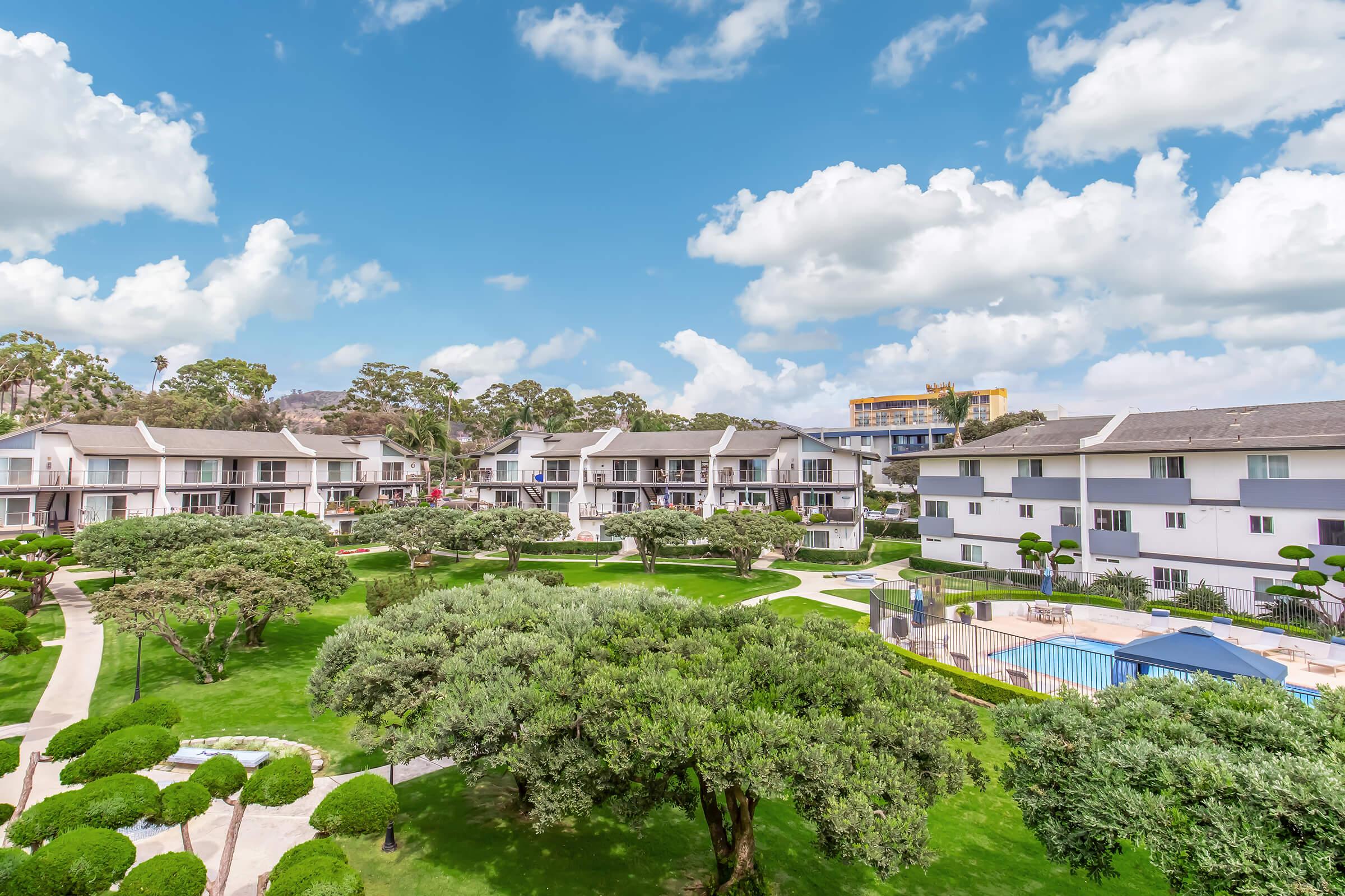 A scenic view of a resort-style courtyard featuring well-maintained green lawns, neatly trimmed shrubs, and several buildings. The sky is bright with fluffy white clouds, and a swimming pool is visible in the area, surrounded by palm trees and lounge chairs. Ideal for relaxation and leisure activities.