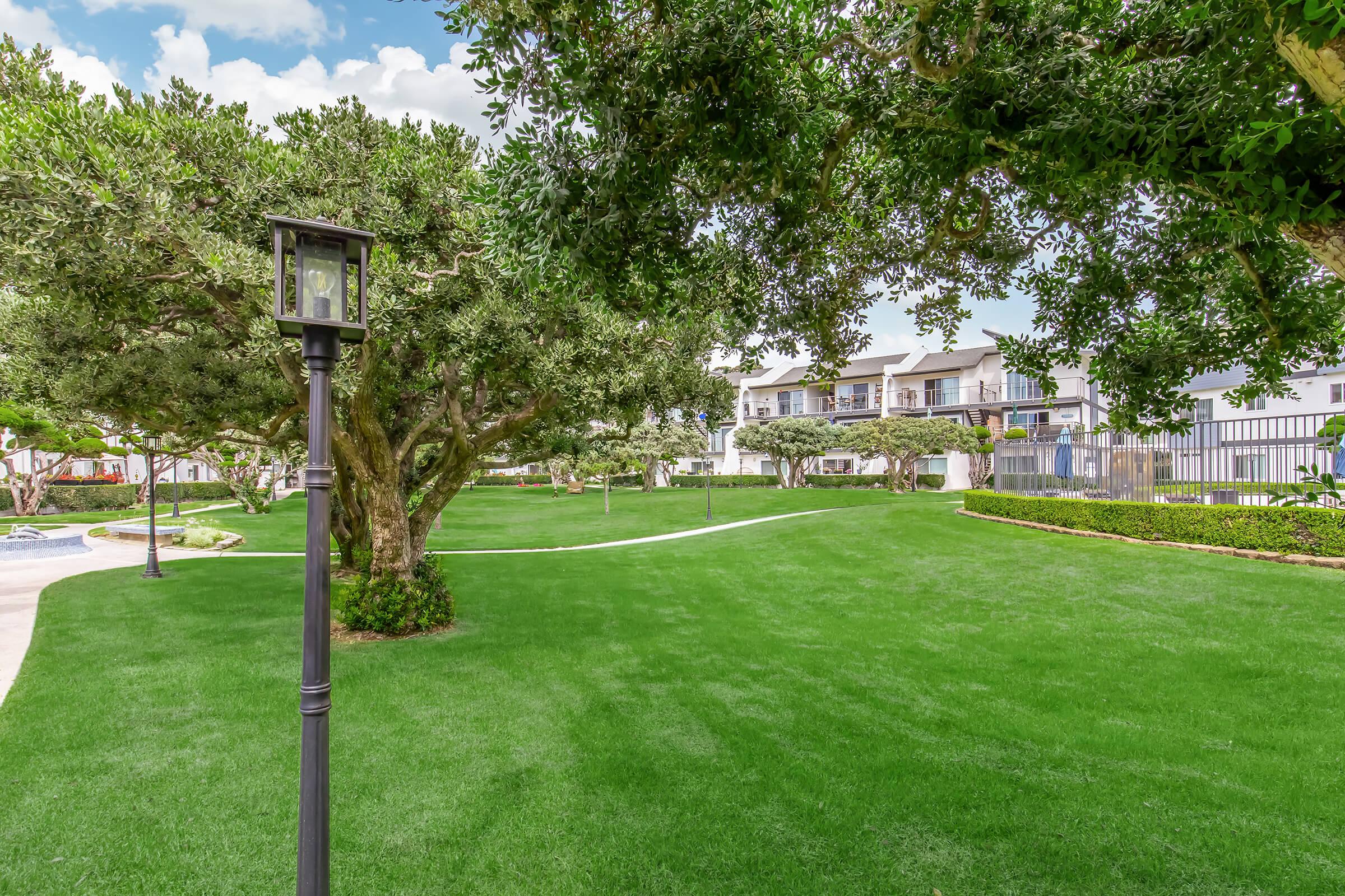 A landscaped outdoor area featuring a manicured green lawn, a vintage-style lamp post, and several trees providing shade. In the background, there are two-story buildings, likely residential, with a pool visible nearby. The scene is sunny and inviting, ideal for relaxation or outdoor activities.