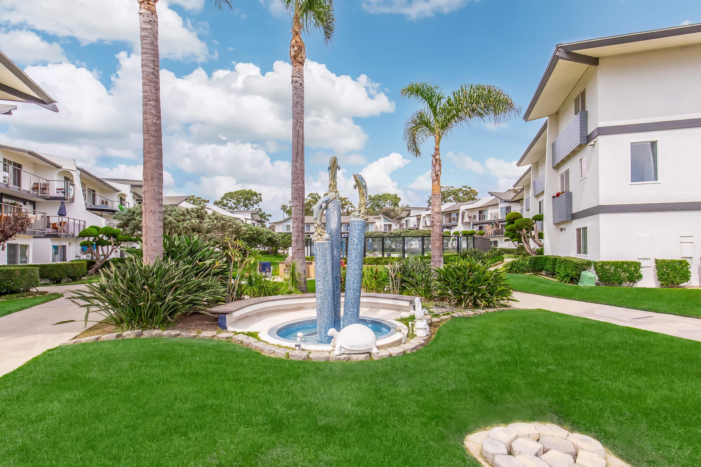 A landscaped courtyard featuring a blue fountain surrounded by greenery and palm trees, with residential buildings in the background. The scene is bright and sunny, showcasing a well-maintained outdoor area with lush grass and decorative plants.