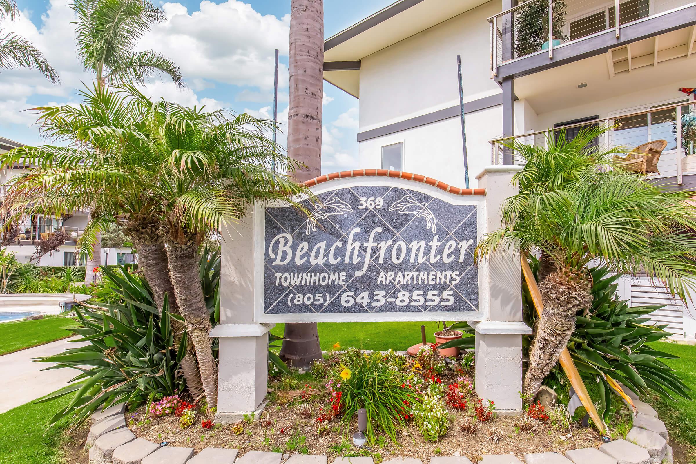 Sign for Beachfronter Townhome Apartments, located at 369, with a contact number (805) 643-8555. The sign is surrounded by tropical plants and palm trees, with a pool visible in the background and a modern building nearby. The sky is partly cloudy.