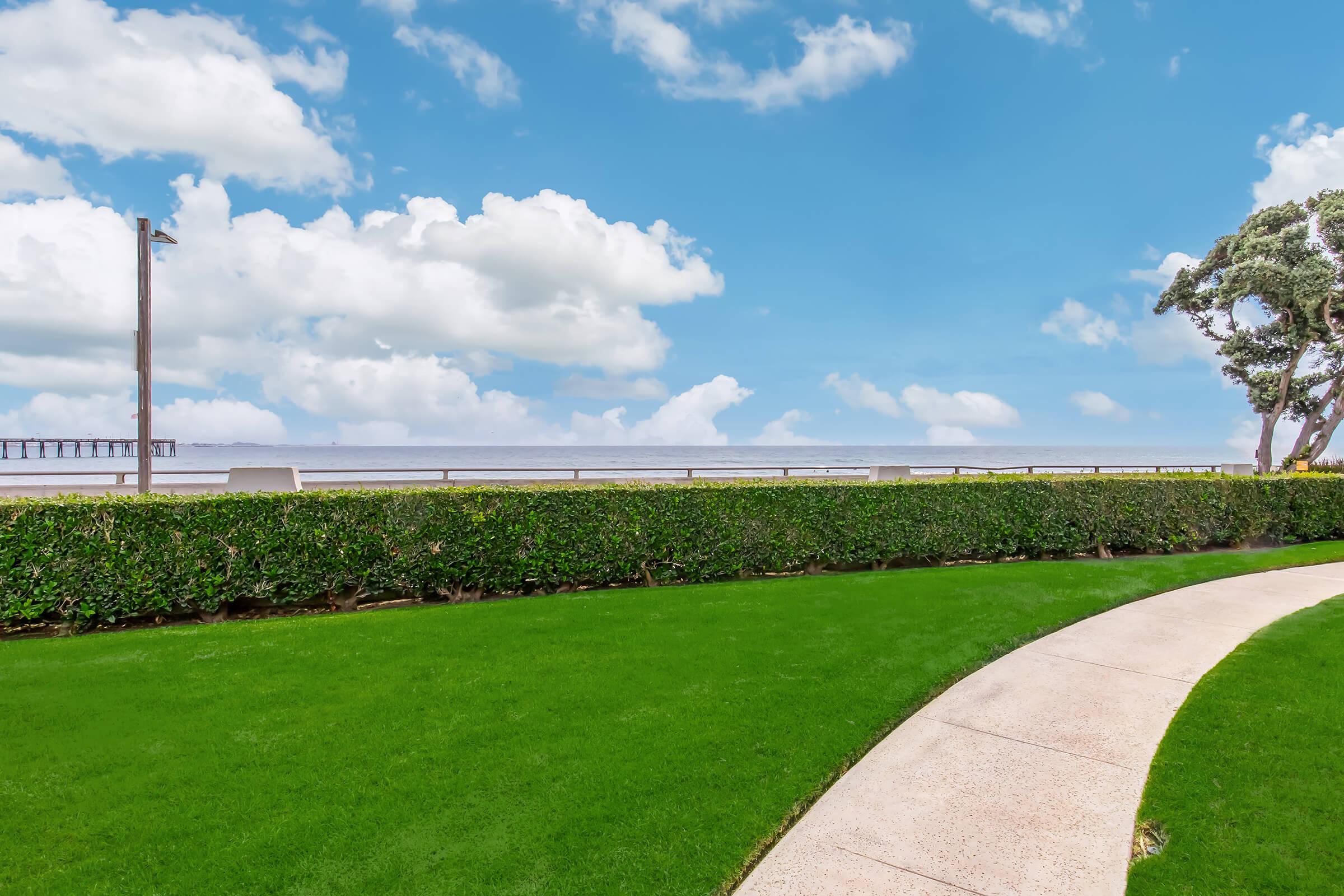 A scenic view of a green lawn bordered by a neatly trimmed hedge, leading to a pathway that curves towards the horizon. In the distance, a calm ocean stretches beneath a bright blue sky with fluffy white clouds, and a pier can be seen against the water.