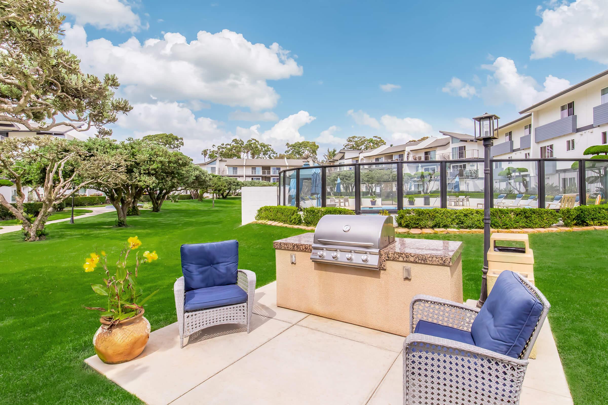 A well-maintained outdoor space featuring a grill, two blue chairs, and a potted plant. The area is surrounded by lush green lawns, trees, and modern residential buildings under a bright blue sky with fluffy white clouds.