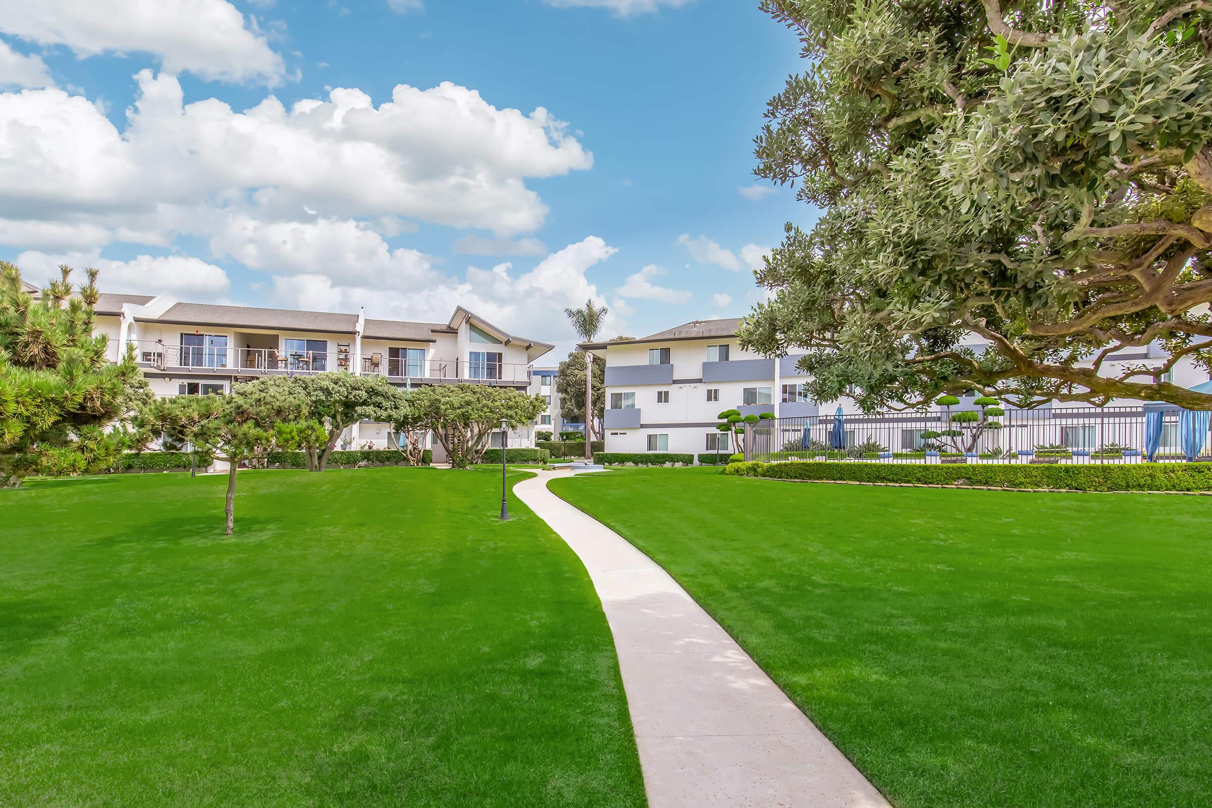 A well-maintained pathway curves through a lush green lawn, leading towards modern white apartment buildings under a bright blue sky with fluffy clouds. Surrounding trees and shrubs enhance the serene landscape of the outdoor area.