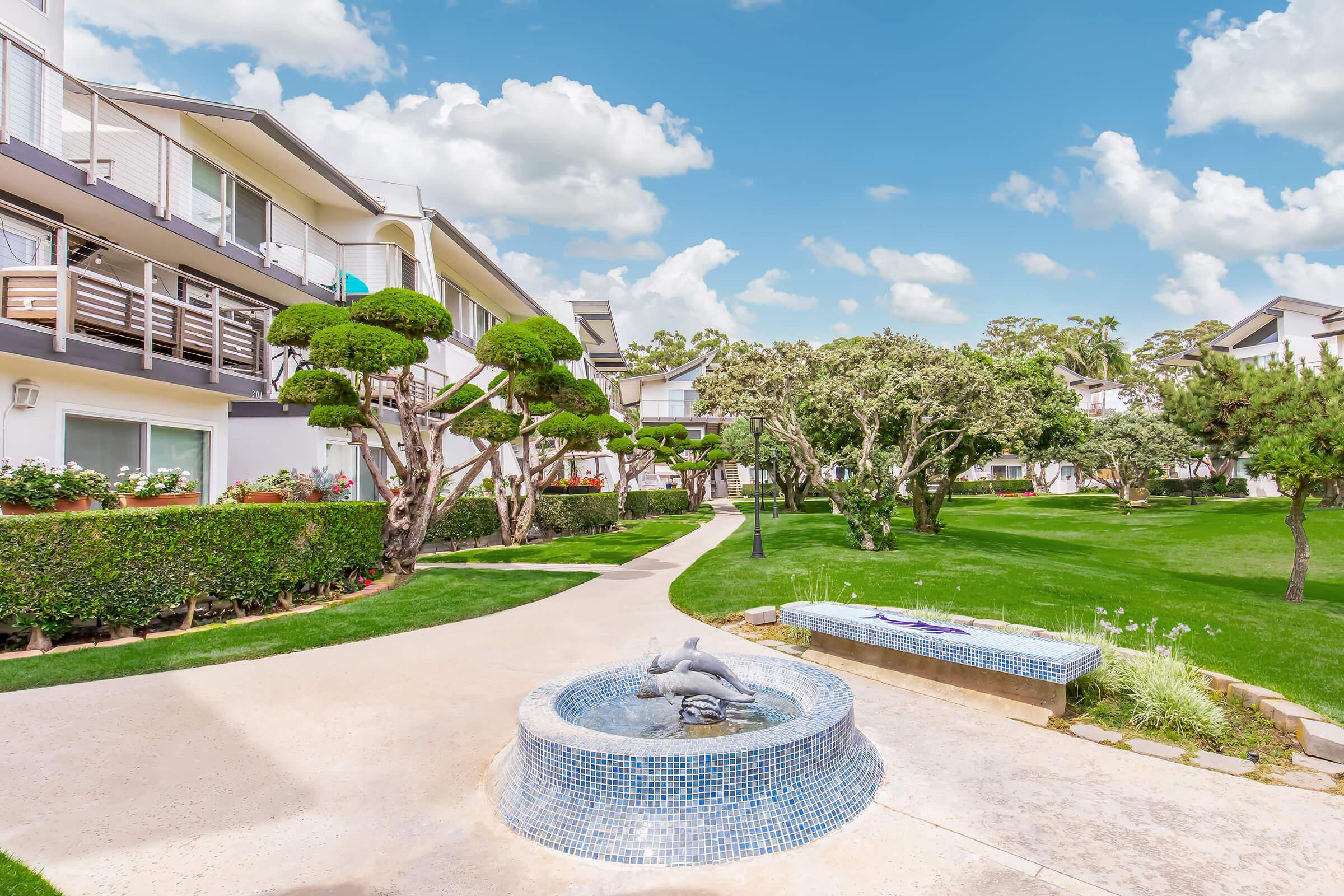 A landscaped courtyard featuring well-manicured grass and trees, with a charming fountain in the center. Surrounding buildings have balconies and flower pots, under a bright blue sky with scattered clouds. Pathways lead through the green spaces, creating a serene atmosphere.
