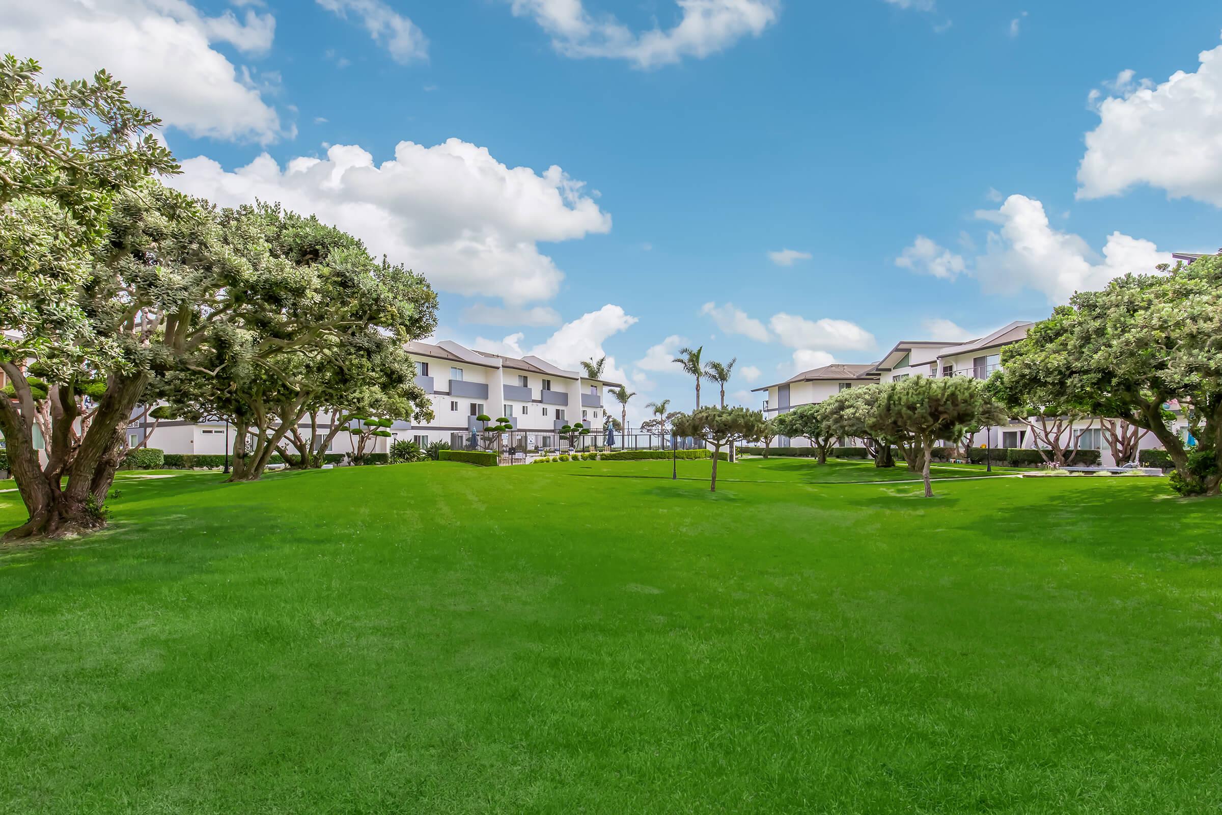 A spacious green lawn with neatly trimmed grass, flanked by trees and shrubs. In the background, modern white buildings with balconies are visible under a bright blue sky dotted with fluffy white clouds. The scene conveys a serene and well-maintained outdoor environment.