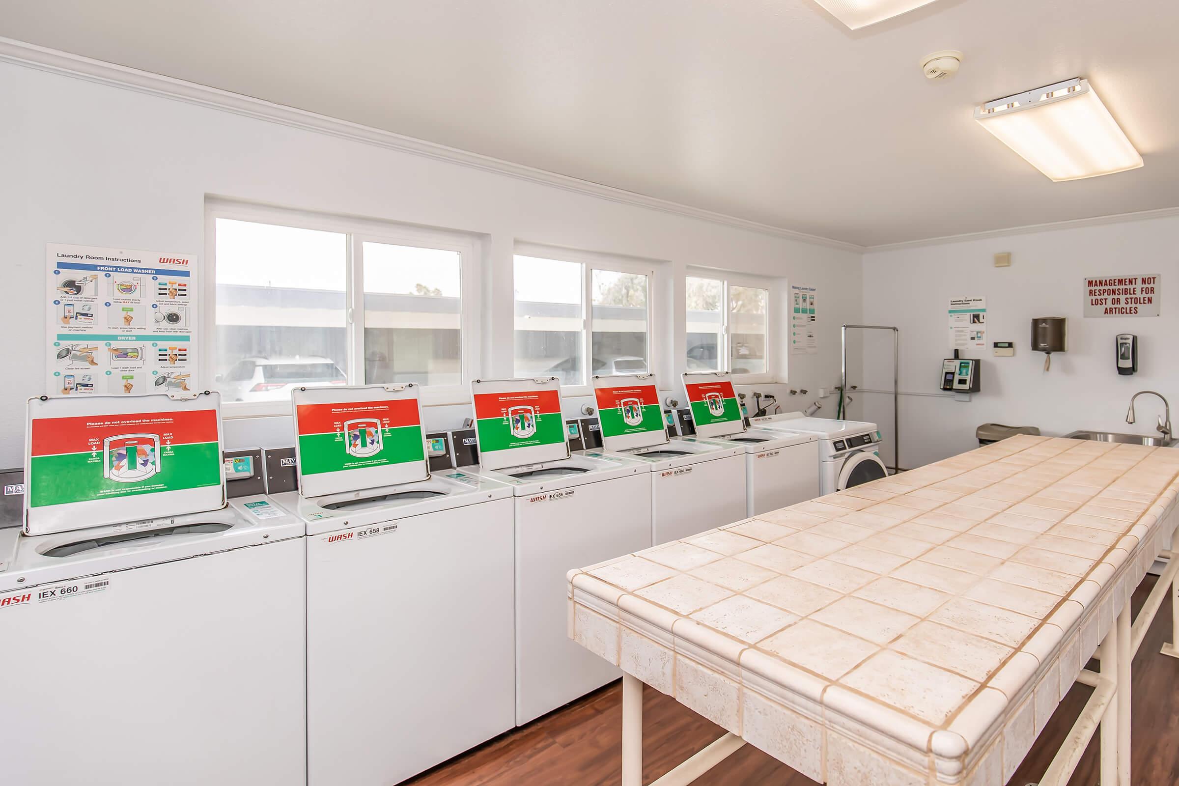 A clean, well-lit laundry room featuring several washing machines with green and red signage on each unit, a large folding table in the center, and windows allowing natural light. Various informational posters about laundry use are visible on the walls.