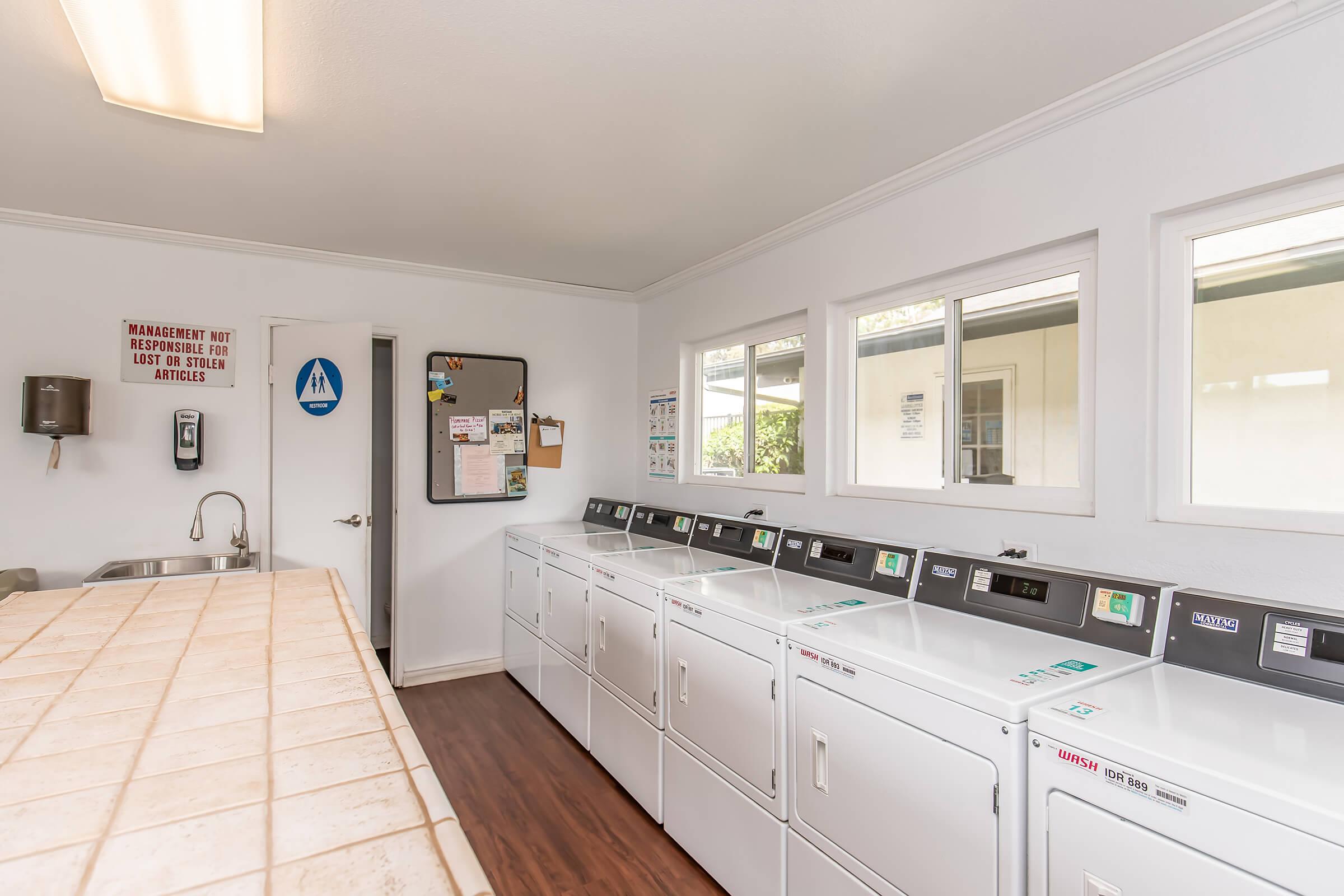 A bright and clean laundry room featuring multiple washing machines and dryers lined up against a white wall. There is a counter in the foreground and a bulletin board on the wall. A door leads to another area, and there are restroom signs visible. The overall atmosphere is tidy and well-maintained.