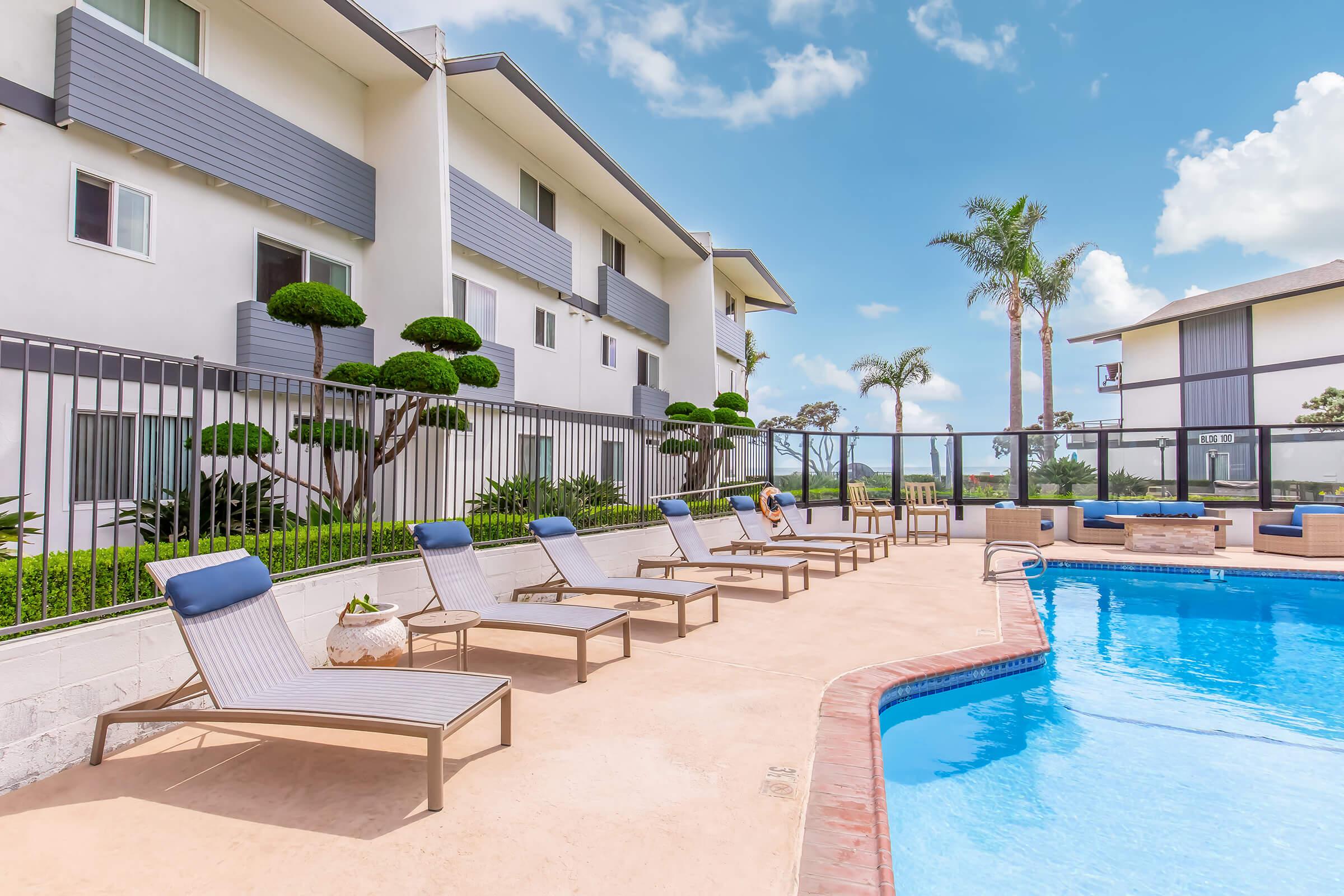 A serene pool area at an apartment complex featuring lounge chairs, manicured shrubs, and palm trees under a bright blue sky. The pool is glistening, with a glass enclosure nearby, creating a relaxing outdoor space for residents.