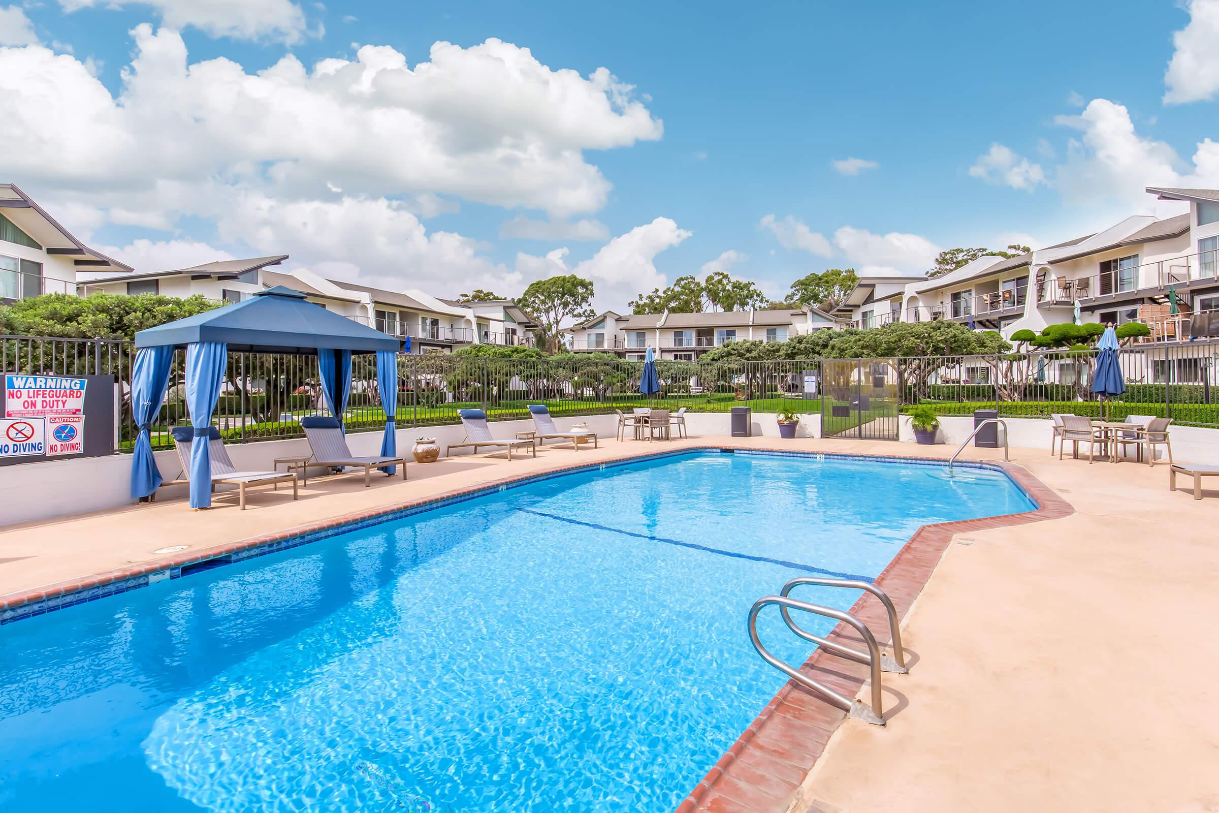 A swimming pool with clear blue water, surrounded by lounge chairs and a shaded gazebo. The background features well-maintained greenery and multi-story residential buildings under a blue sky with fluffy clouds.