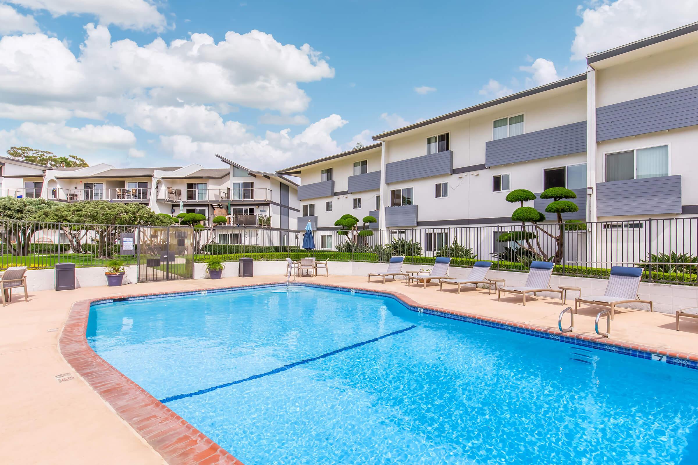 A clear blue swimming pool surrounded by lounge chairs, with landscaped trees and shrubs. In the background, modern apartment buildings feature balconies and gray siding under a bright blue sky with fluffy clouds.