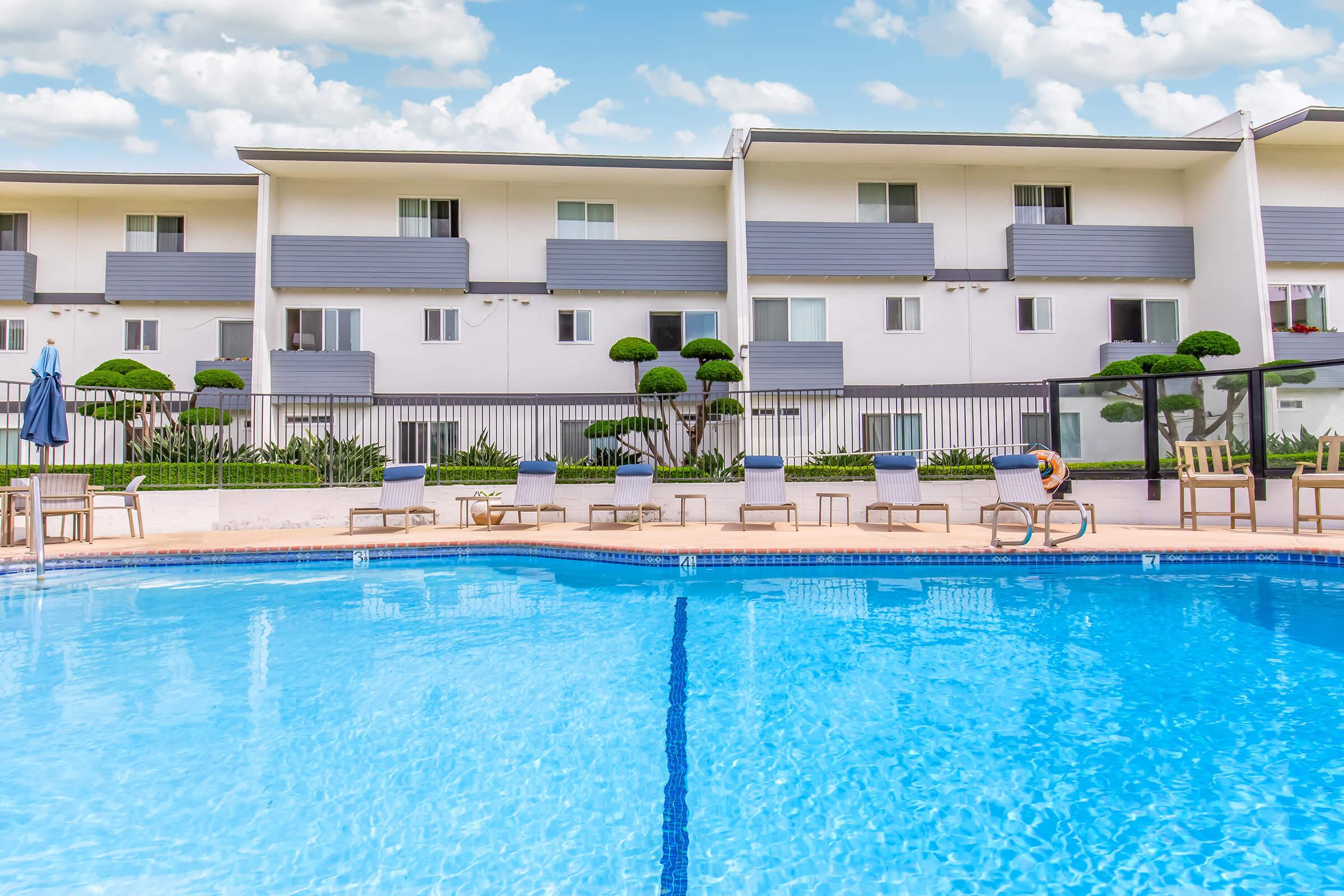 A clear blue swimming pool surrounded by lounge chairs. In the background, there are modern buildings with gray siding and manicured shrubs. The sky above is bright with fluffy white clouds.