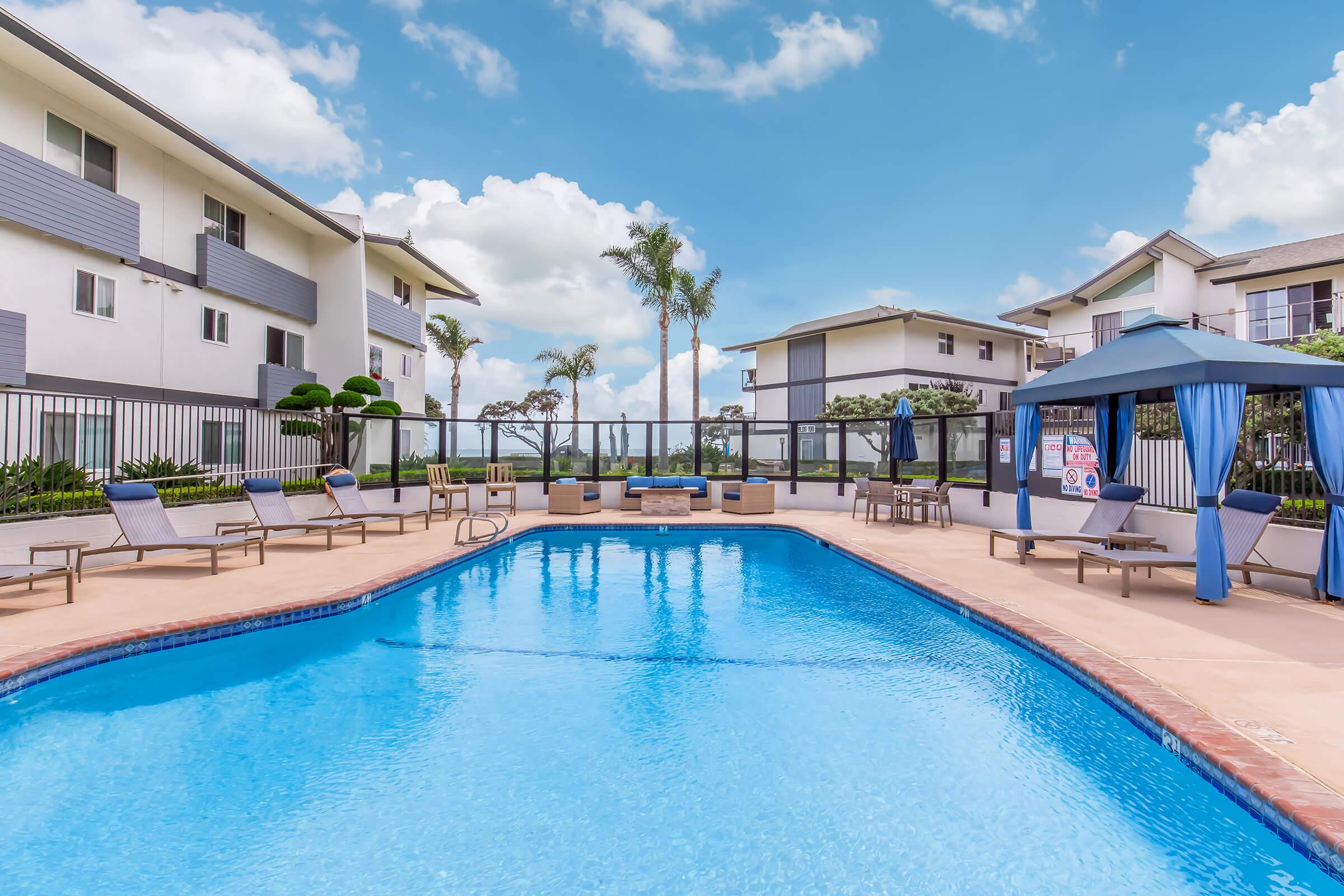 A serene outdoor pool area surrounded by palm trees and lounge chairs. The pool features a blue water surface under a clear sky with fluffy clouds. In the background, modern apartment buildings are visible, along with a gazebo providing shaded seating.