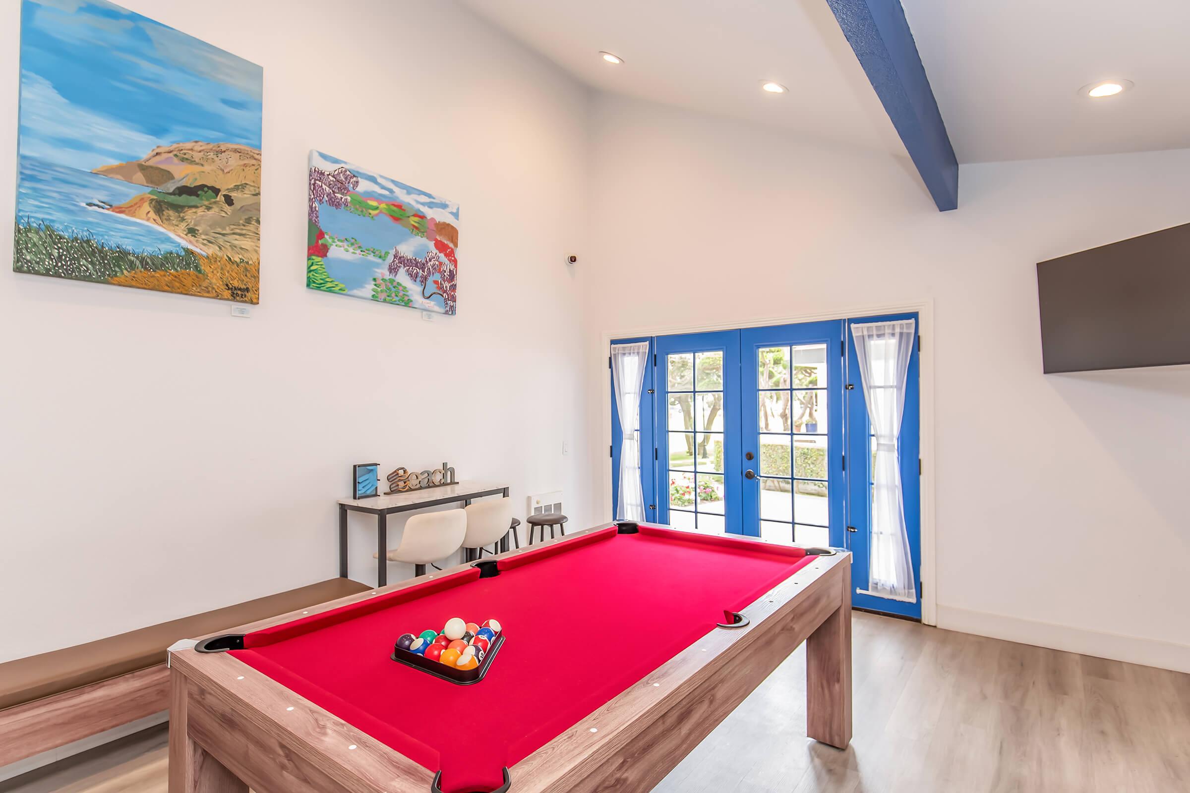 A well-lit game room featuring a red pool table with balls arranged in a triangle, two colorful landscape paintings on the wall, and large blue French doors leading to an outdoor area. A modern chair and a small decorative table are visible, enhancing the inviting atmosphere.
