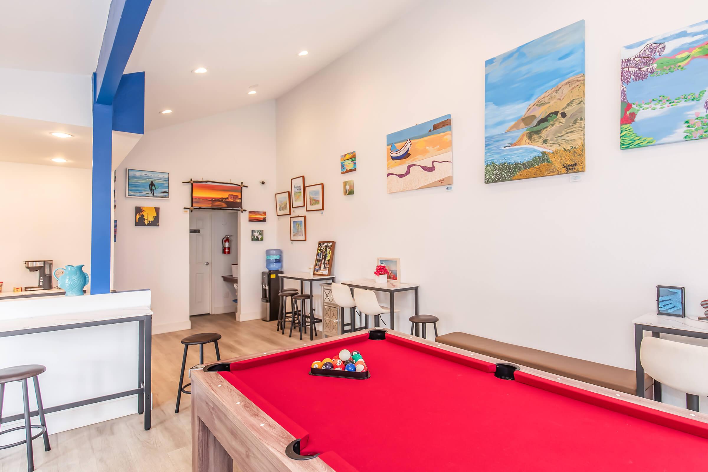 A bright interior of a recreation room featuring a red pool table with balls arranged on it. The walls are adorned with colorful artwork, and there are simple bar stools and small tables nearby. The space has large windows and a welcoming atmosphere, perfect for socializing and enjoying leisure activities.