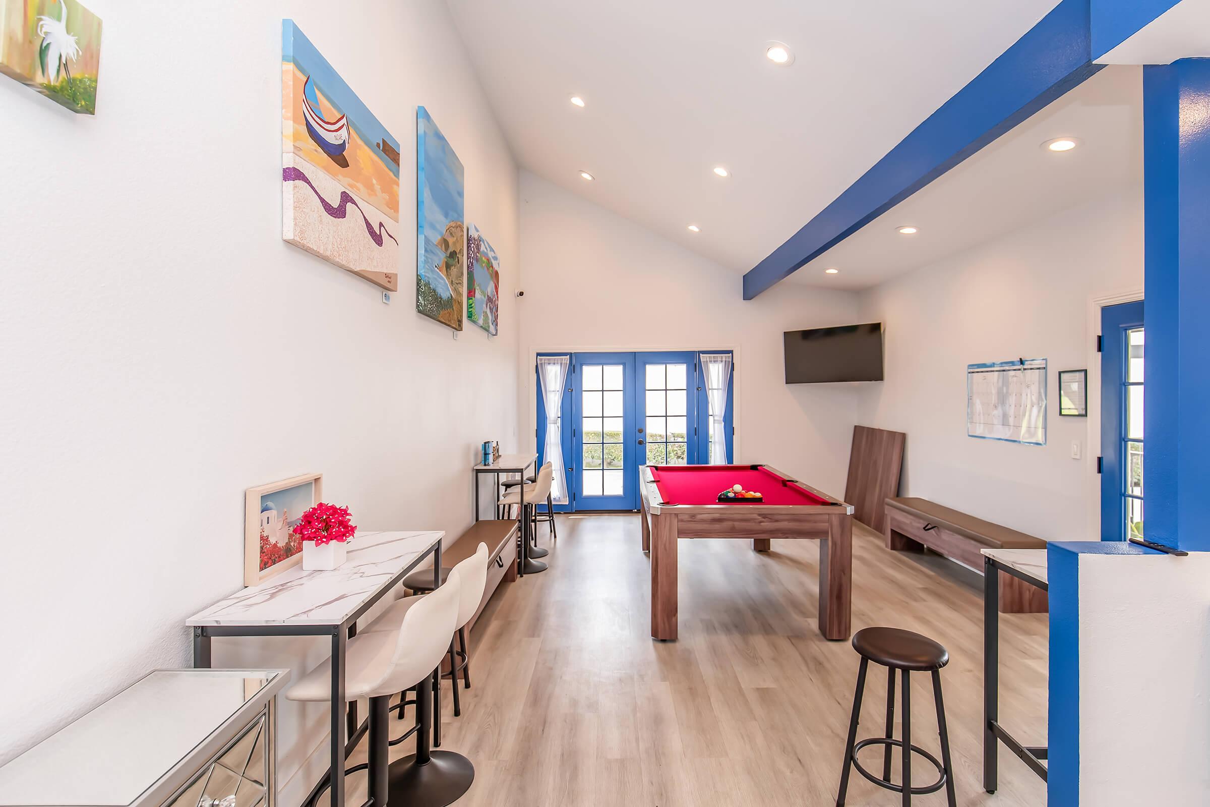 A bright and airy room featuring a red pool table, sleek bar stools, and a dining area. The walls are adorned with colorful artwork, and large windows allow natural light to flood the space. A flat-screen TV is mounted on one wall, adding to the modern, inviting atmosphere.