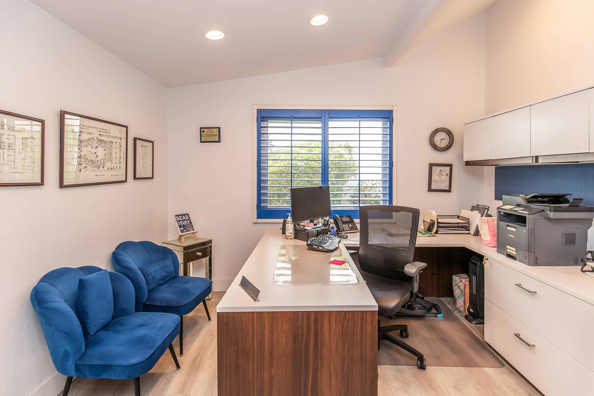 A modern office space featuring a desk with a computer, printer, and office supplies. There are two blue chairs in front of the desk, a small side table, and wall decorations including framed documents. Natural light enters through a window with blue blinds, enhancing the bright and organized atmosphere.