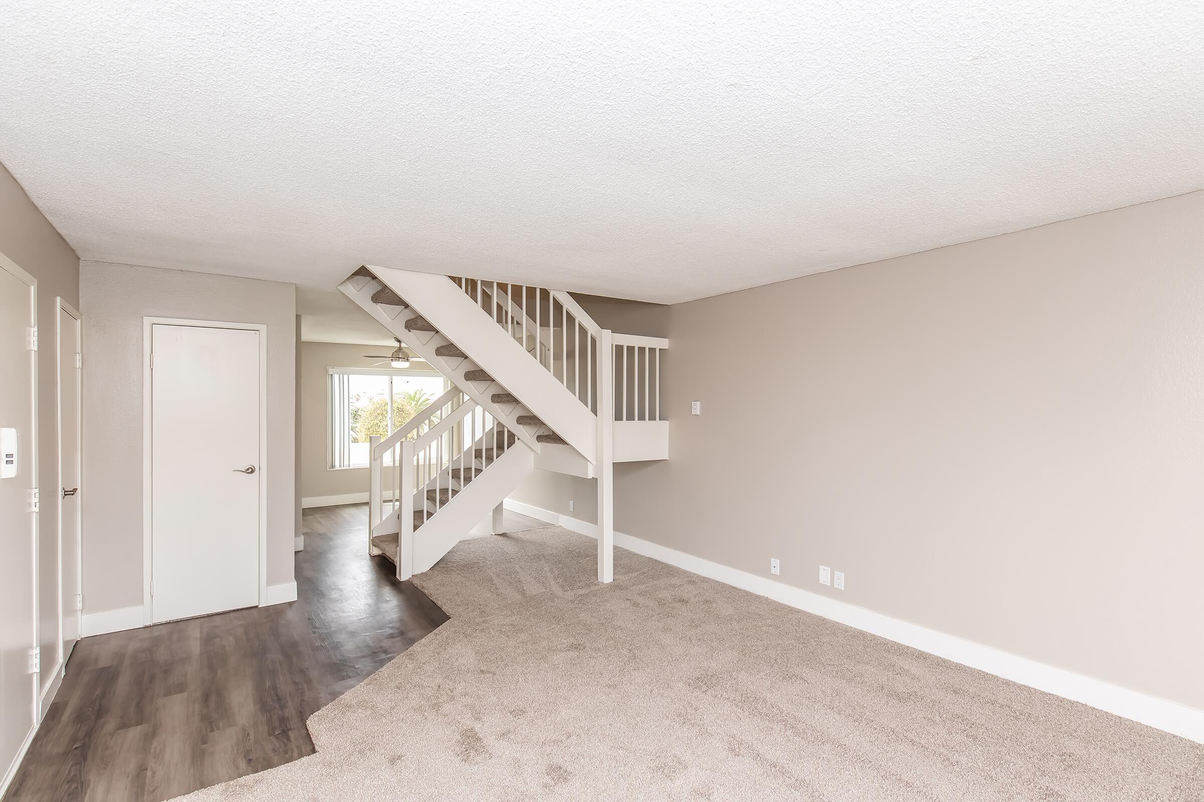 Spacious interior of a room featuring light gray walls, a carpeted floor, and a staircase leading to an upper level. A large window offers natural light, and a white door is visible on the left. The space appears inviting and modern, suitable for various layouts and furnishings.