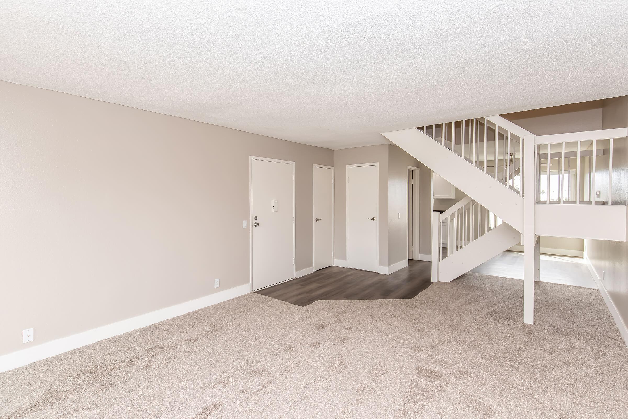 A spacious, empty room featuring light beige walls and a plush carpet. There are two doorways leading to adjacent rooms, and a staircase with a white railing in the background, creating a bright and open living space.
